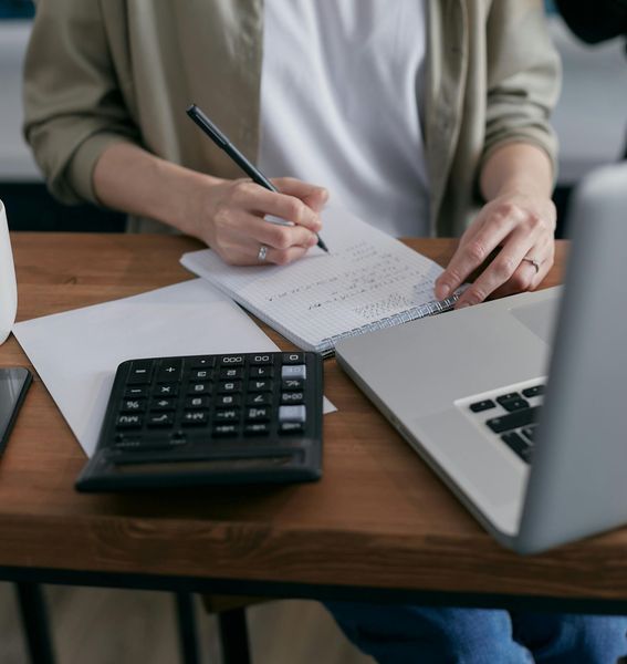 Person writing in a notebook at a desk with a calculator and laptop.