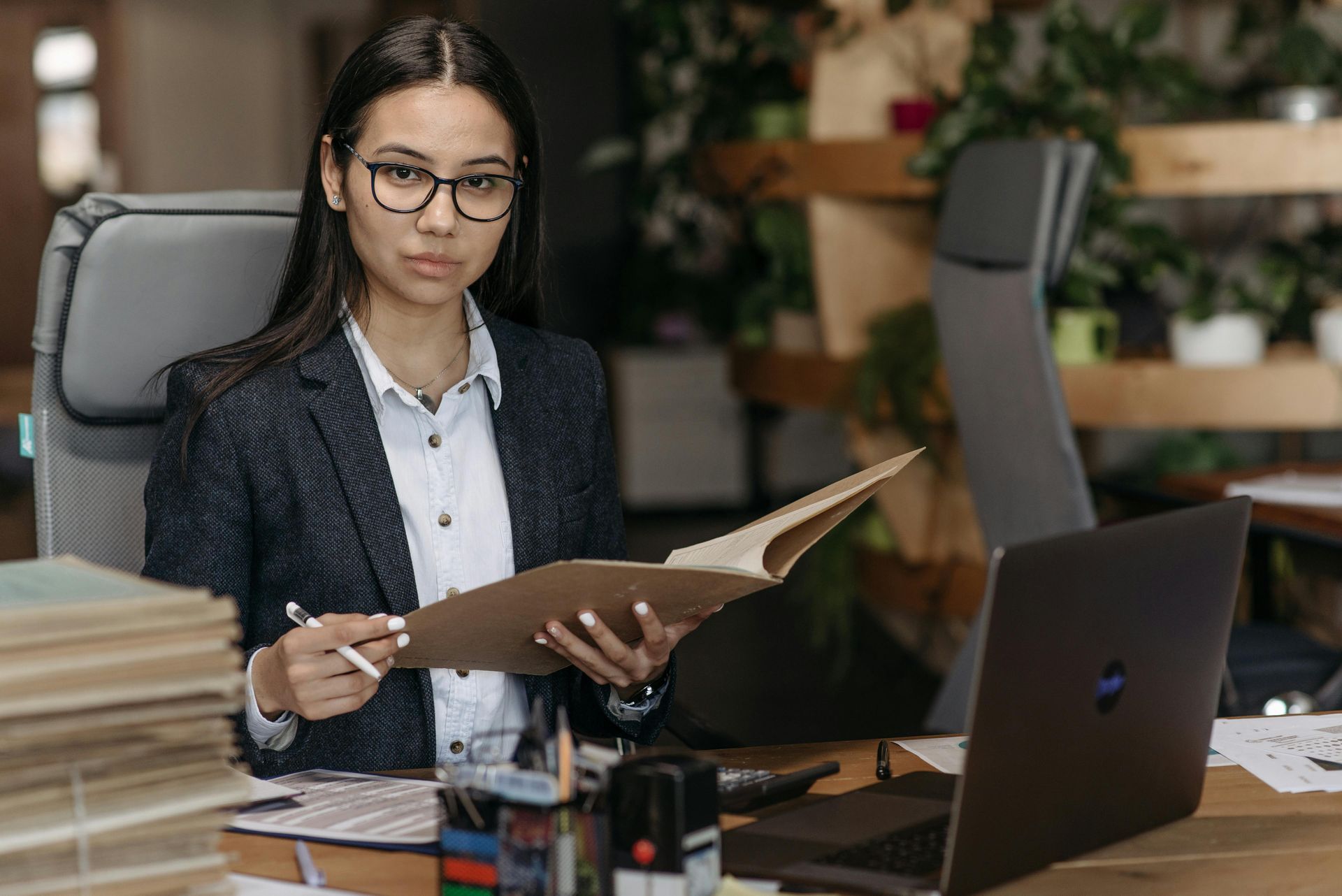 Woman in glasses and blazer at desk, holding a folder, looking at the camera.