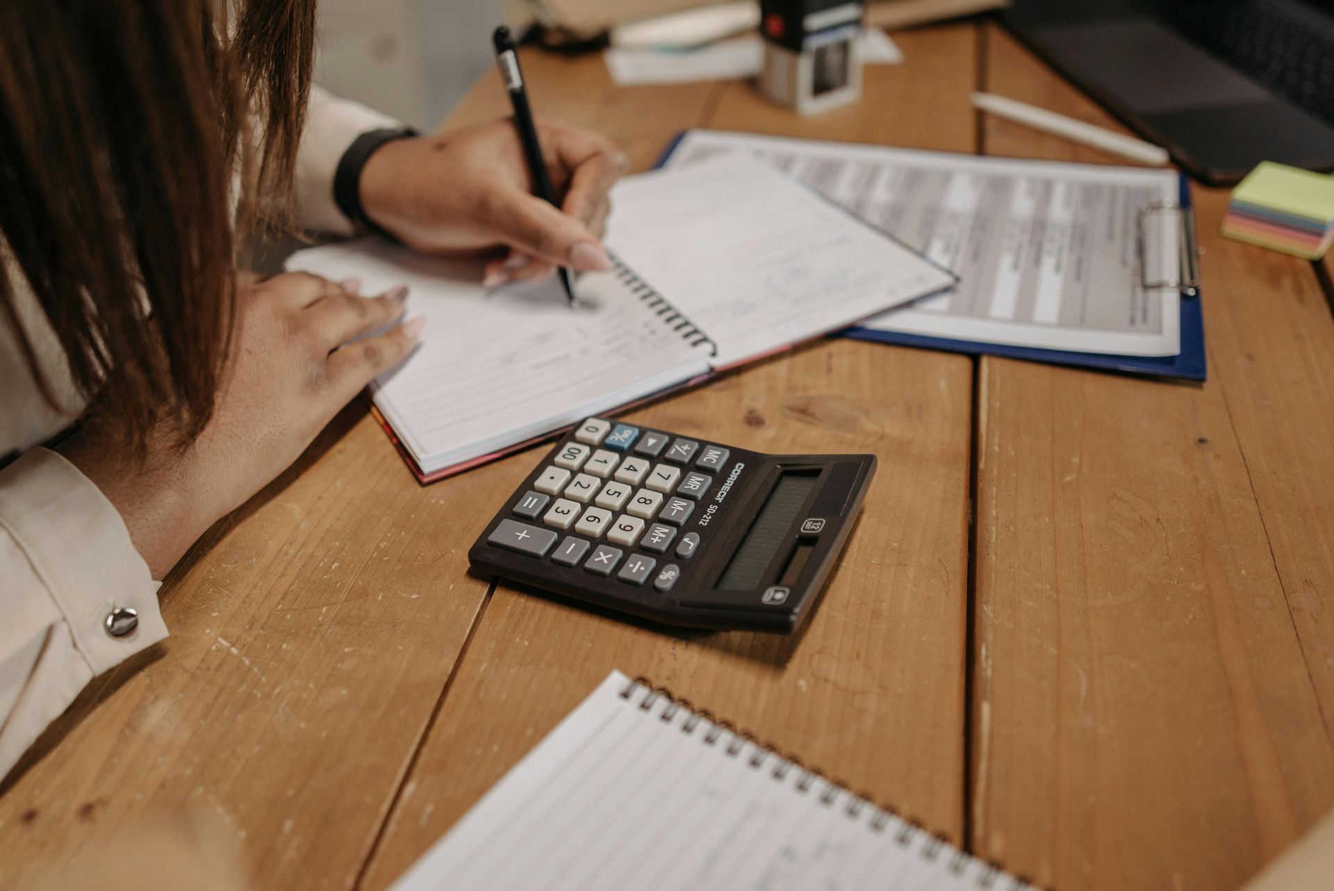 Person writing in a notebook, using a calculator on a wooden desk with other papers.