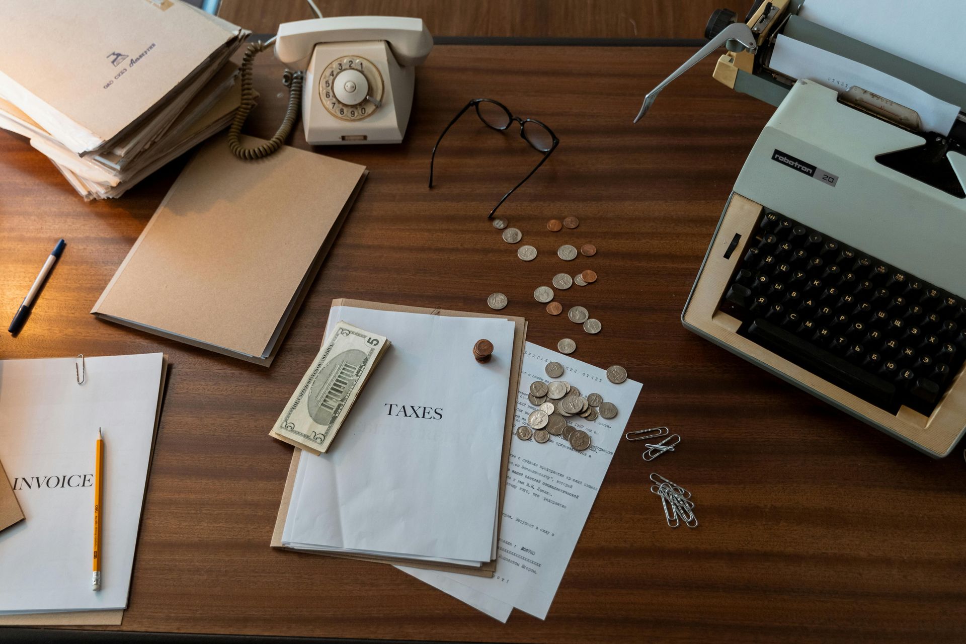 Desk with a typewriter, rotary phone, documents, money, and glasses.