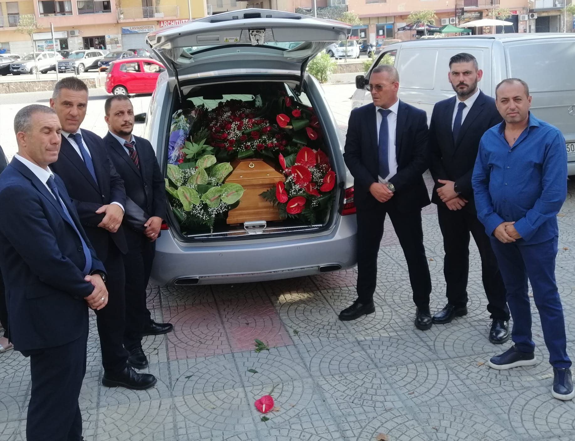 Uomini in giacca e cravatta sono in piedi vicino a un carro funebre con la parte posteriore aperta, contenente una bara e composizioni floreali.