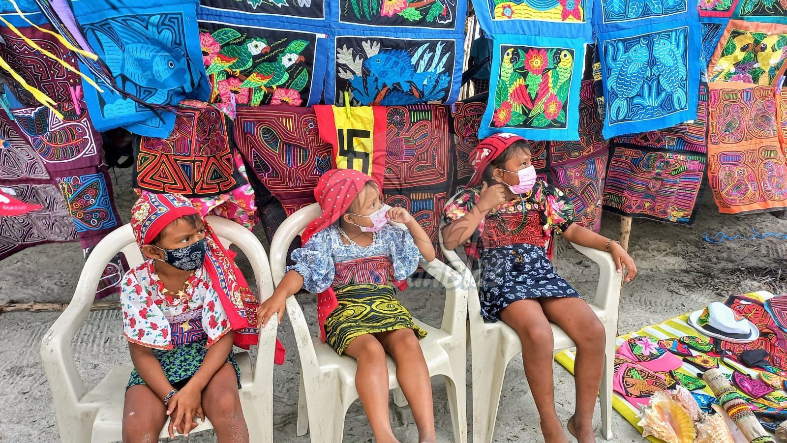 three girls wearing face masks sit in chairs in front of a display of colorful fabrics