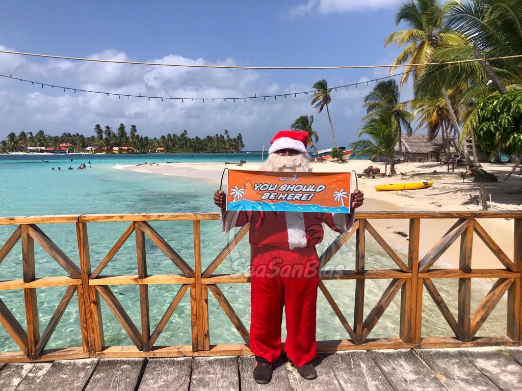 A man dressed as santa claus is holding a sign in front of a beach.