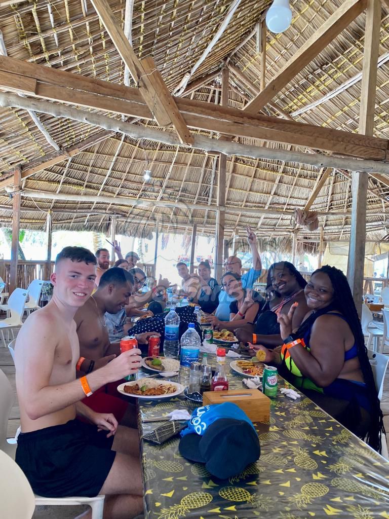 A group of people are sitting at a table eating food under a thatched roof.