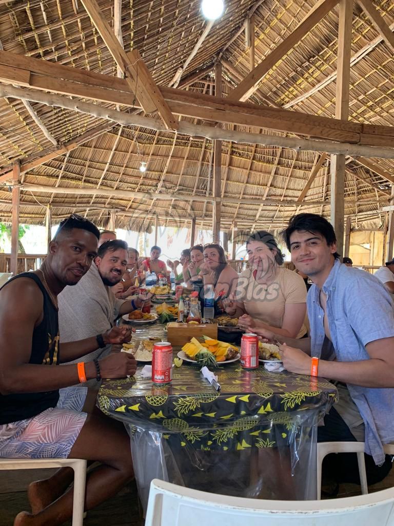 A group of people are sitting at a table eating food under a thatched roof. San Blas Islands Guna Yala San Blas Tours