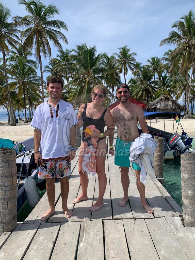 Three people are standing on a wooden dock near a beach.