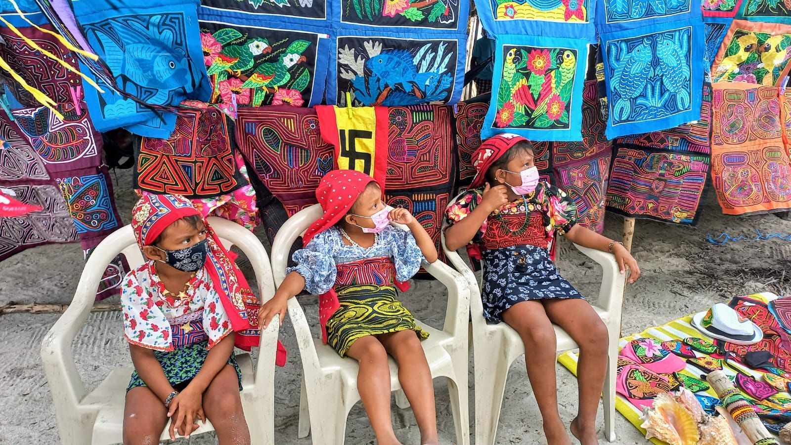 Three young girls wearing masks are sitting in chairs at a market on a Panama San Blas Tour