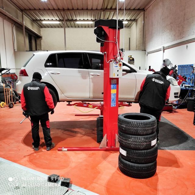 Due meccanici lavorano su un'auto bianca in un'officina con il pavimento rosso. Gli pneumatici sono ammucchiati lì vicino.