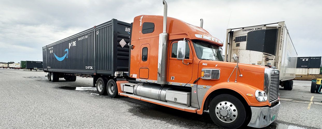 Orange semi-truck with a black Amazon trailer parked on a gray surface under a cloudy sky.