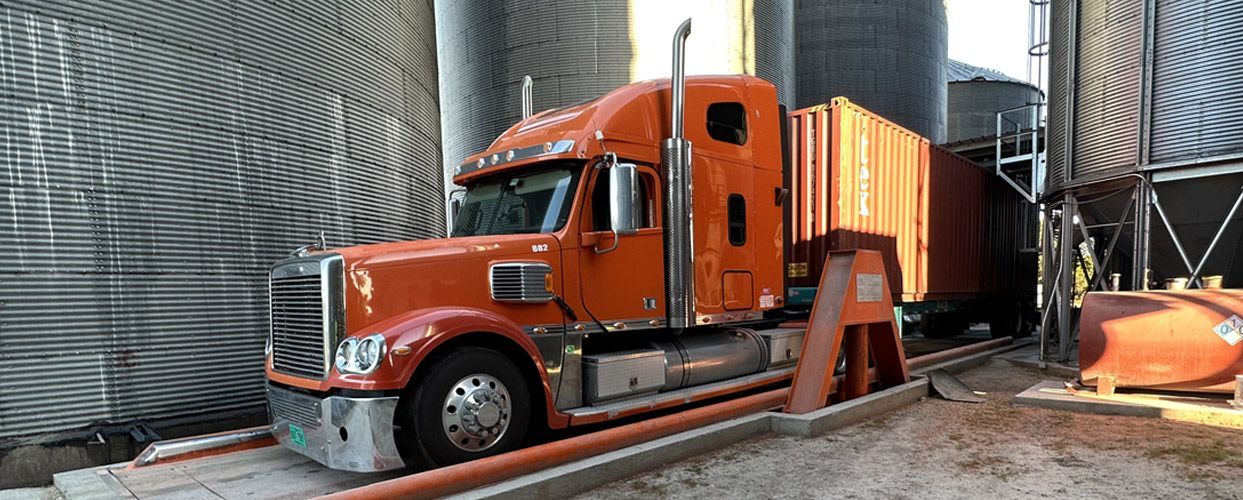 Orange semi-truck loaded with a container entering a grain storage facility.