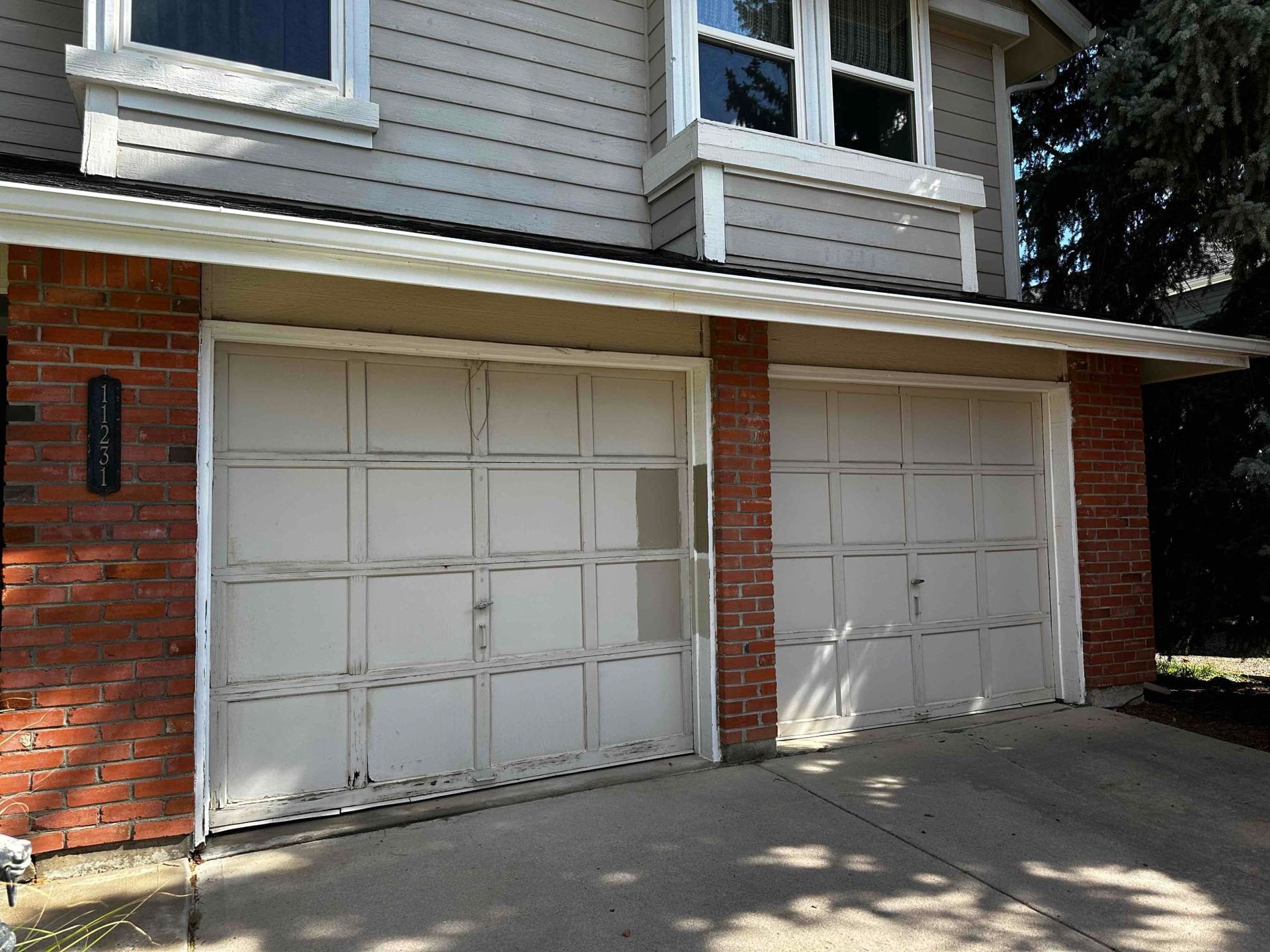 Two-car garage with beige doors and brick columns, set beneath a two-story house with gray siding.