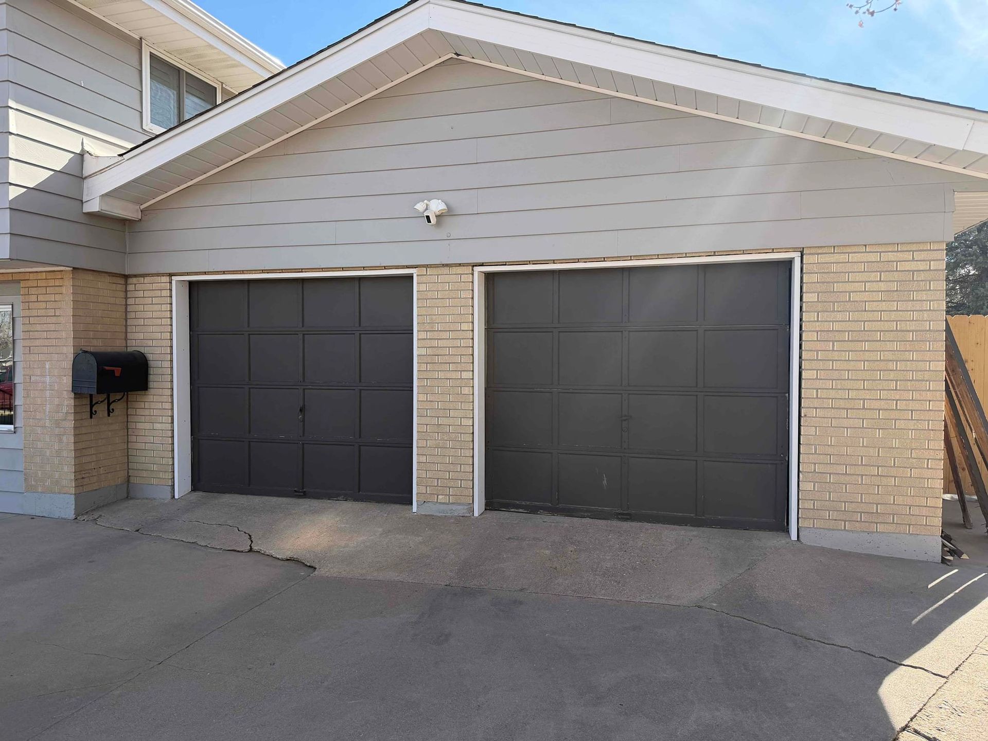 Two-car garage with dark gray doors and light beige brick exterior. Concrete driveway and mailbox visible.