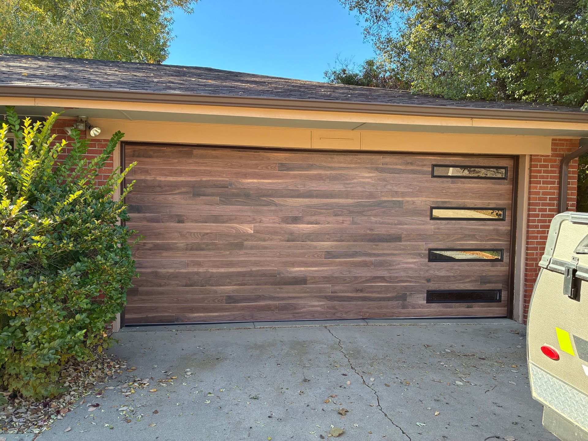 Brown garage door with rectangular windows, brick exterior, and a small green bush.