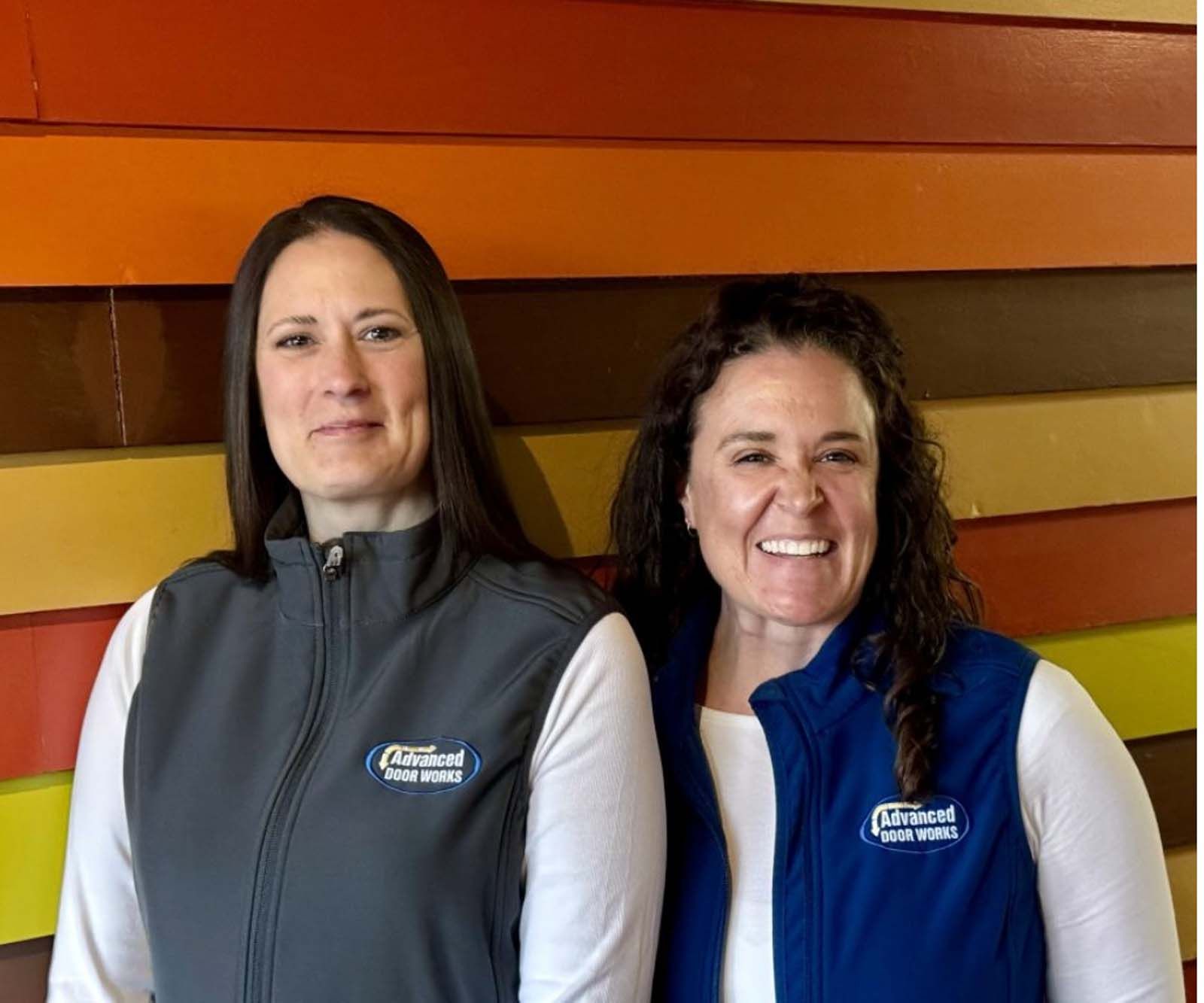 Two women wearing vests are standing next to each other in front of a colorful wall.