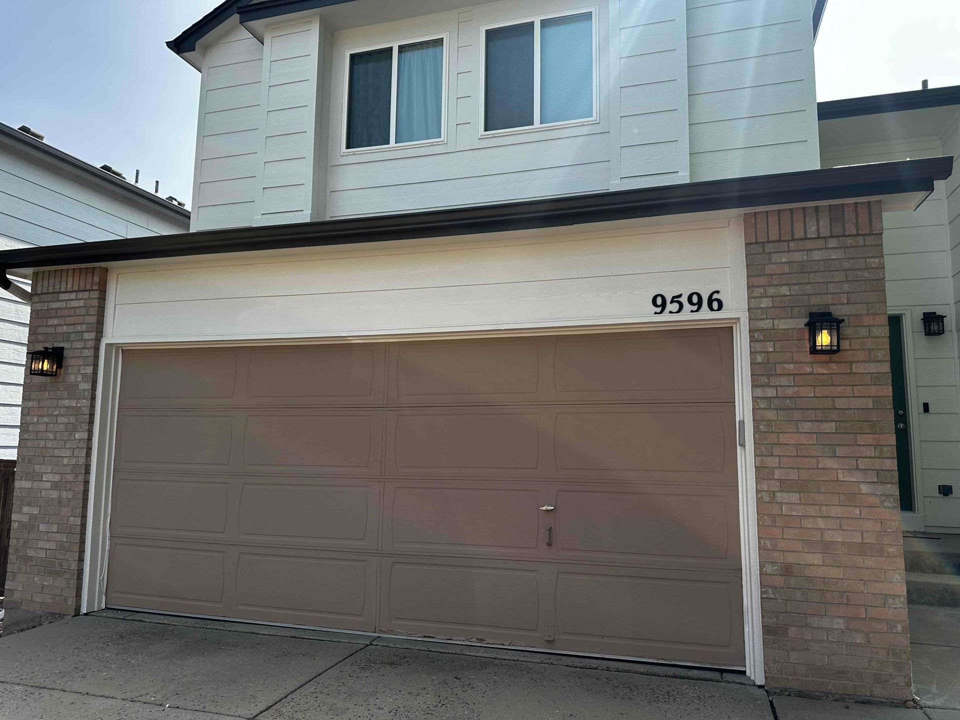 A white house with a brown garage door and a brick wall.