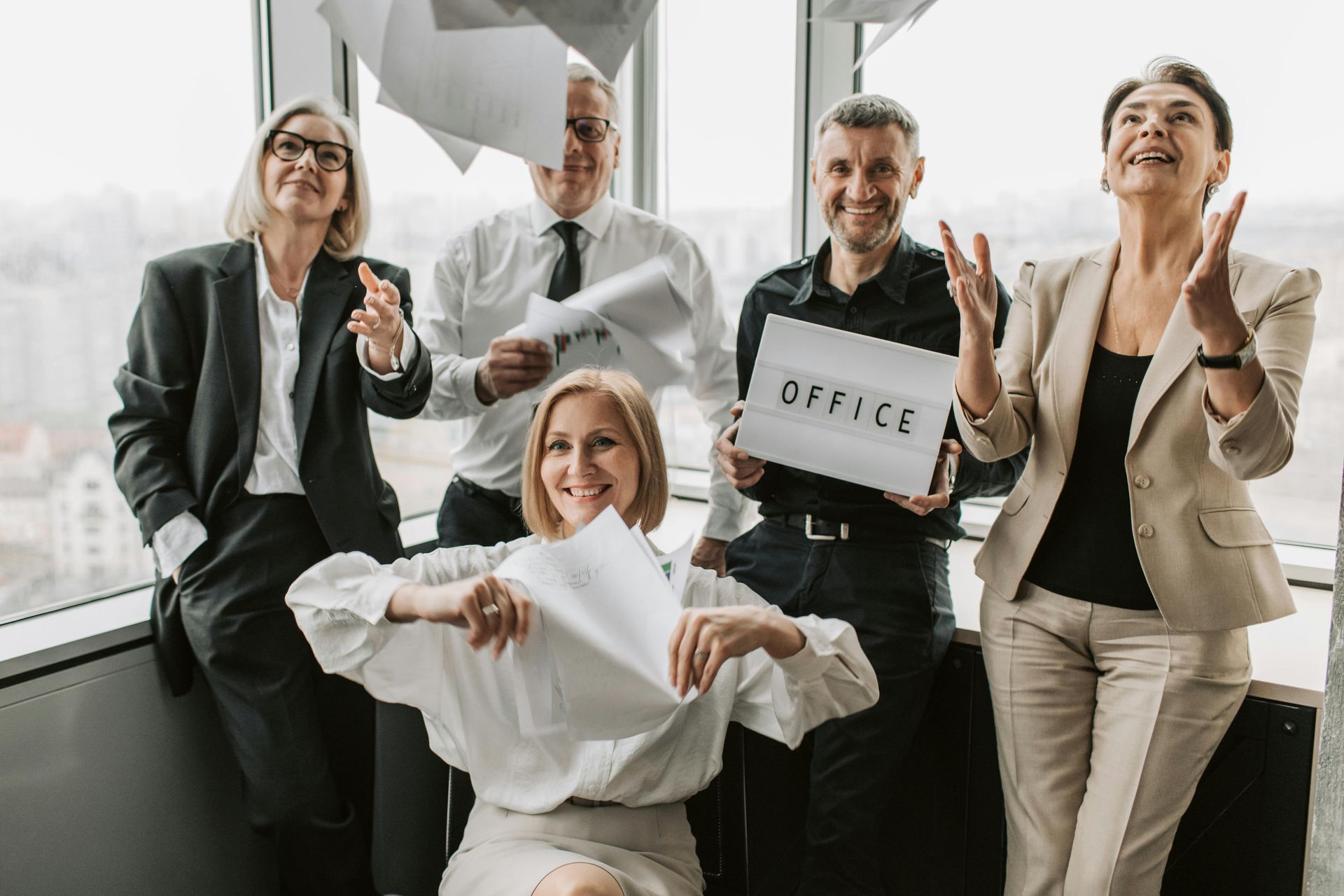 A group of business people are standing in front of a window holding papers and a sign that says office.