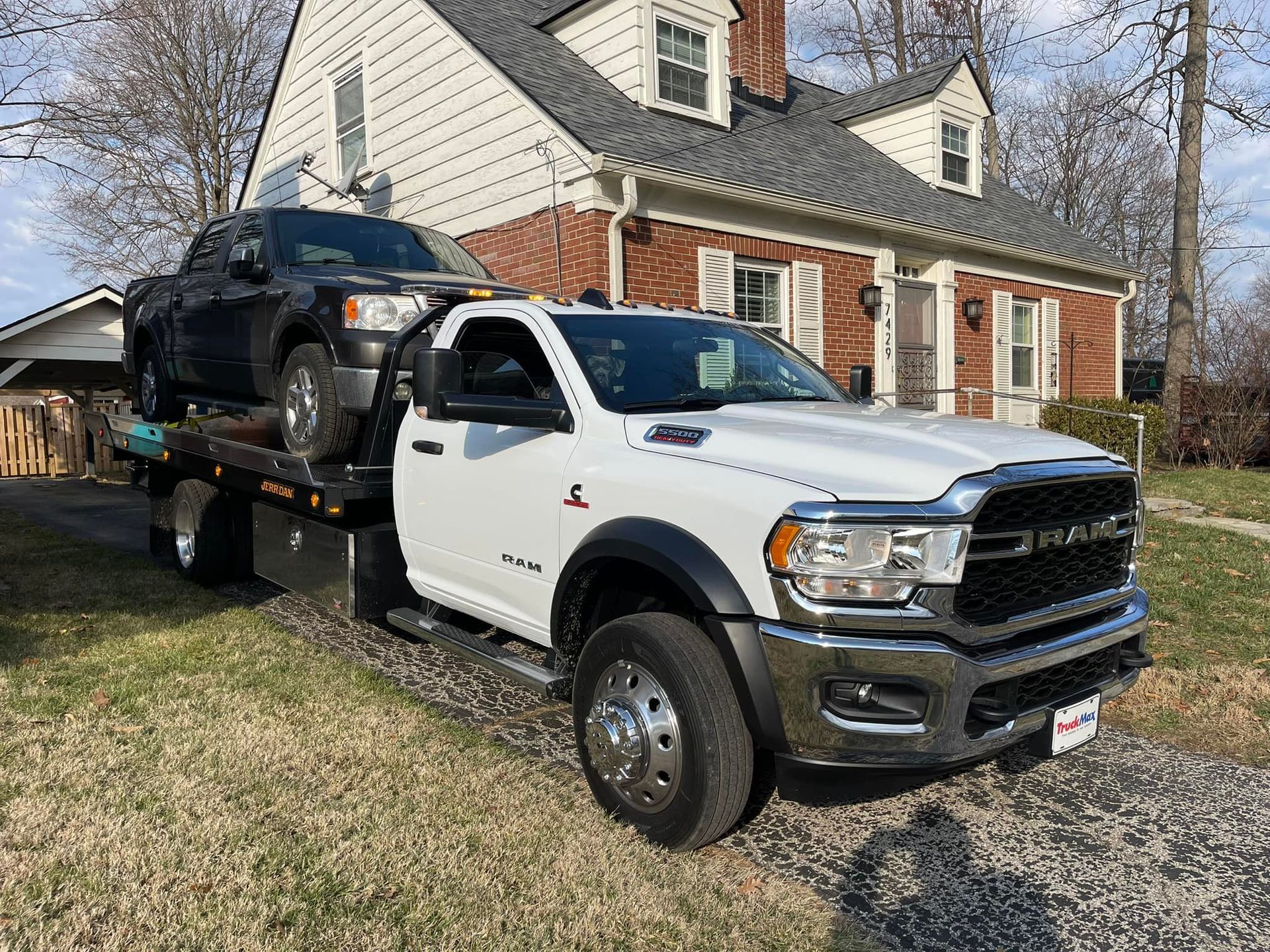 White tow truck with a black pickup truck loaded, in front of a house.
