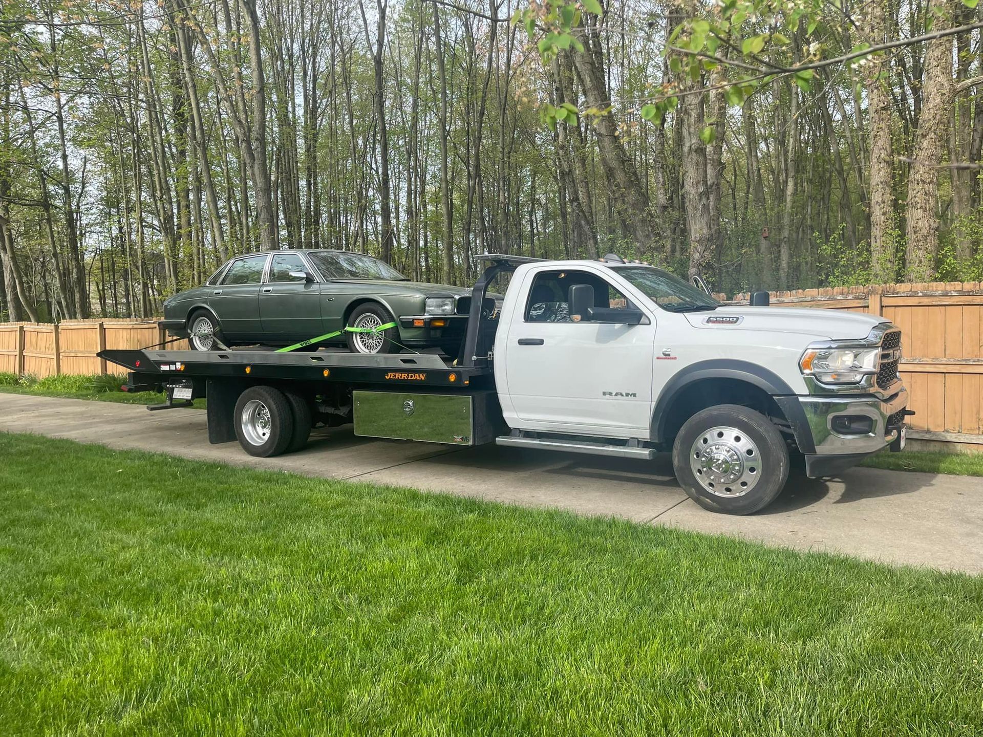 A flatbed tow truck, white, carrying a green car on grass in front of a fence and trees.