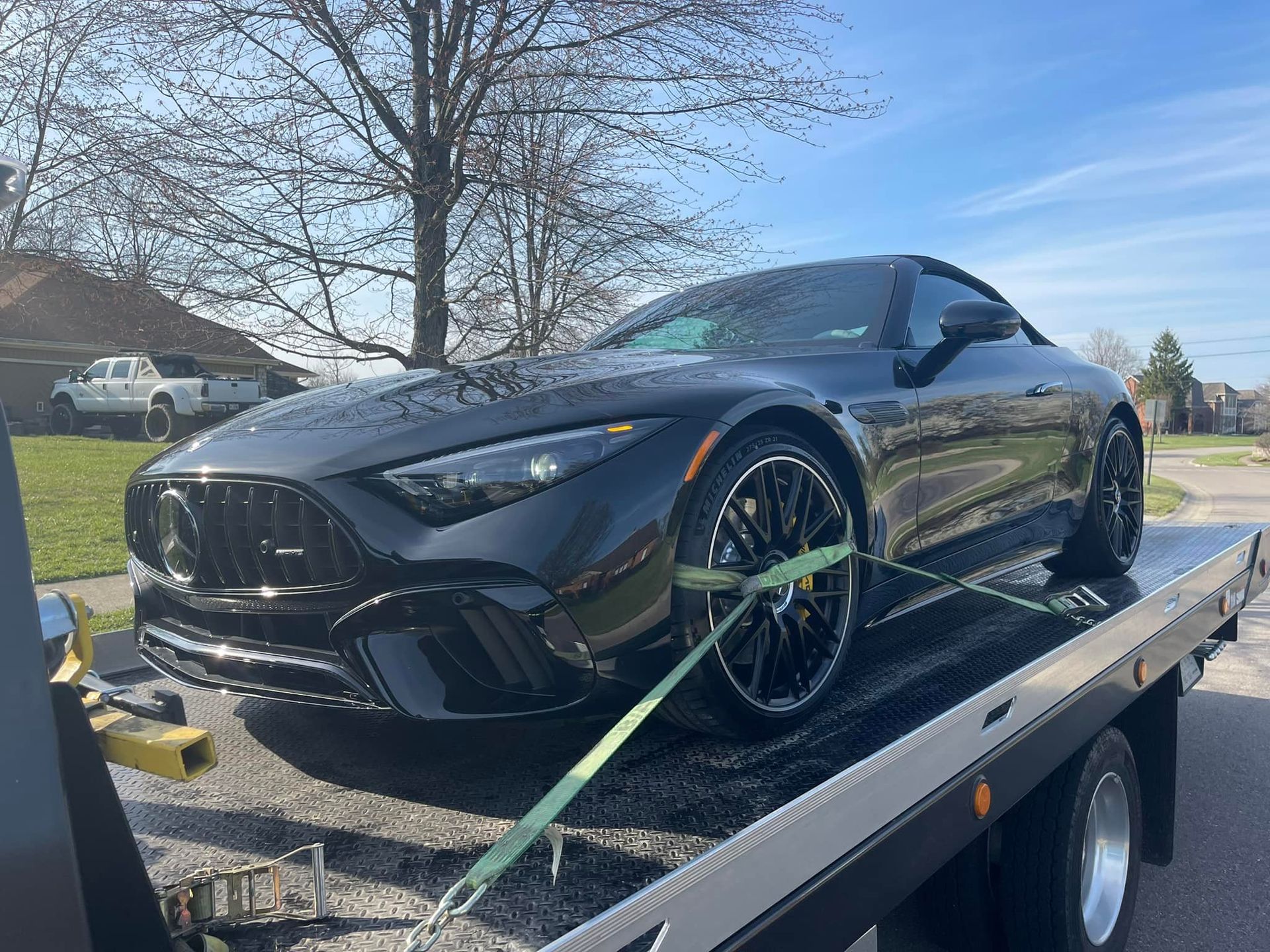 Black Mercedes AMG SL on a flatbed tow truck, secured with straps. Sunny day.