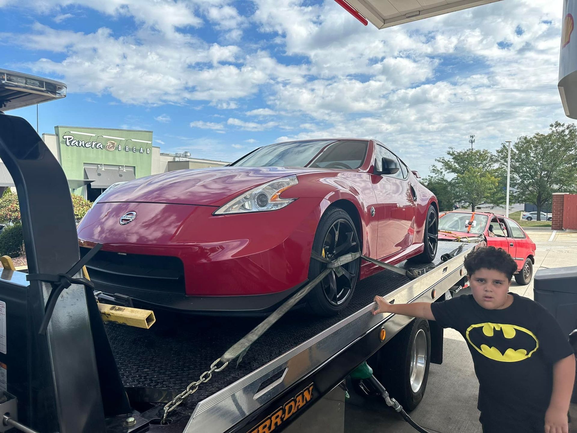 Red Nissan 370Z on a tow truck with a child wearing a Batman shirt.