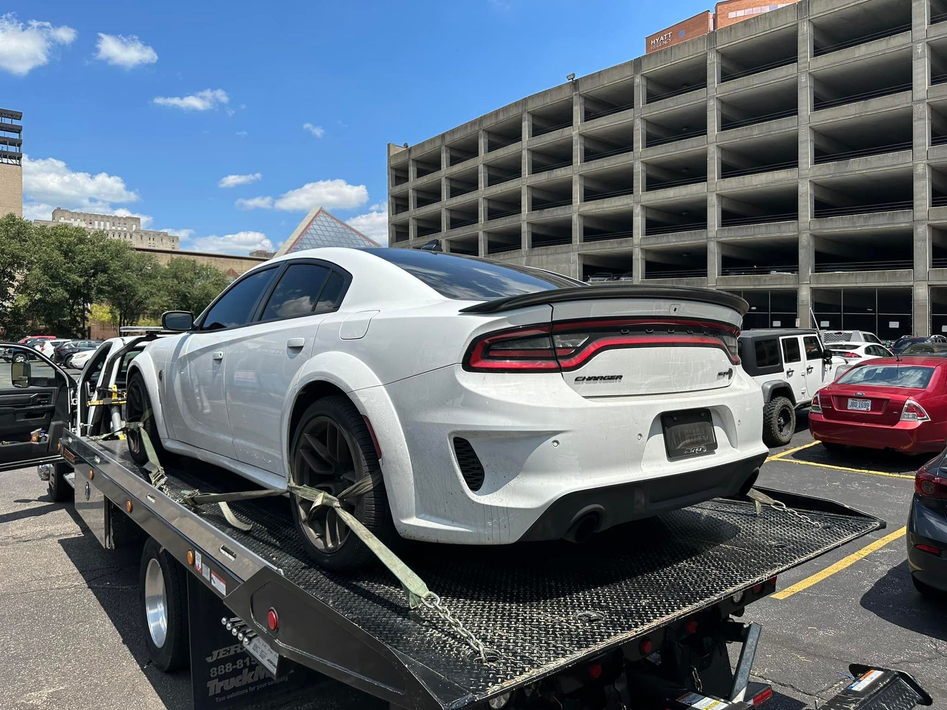 White Dodge Charger on a tow truck, outdoors near a parking garage. Sunny day.
