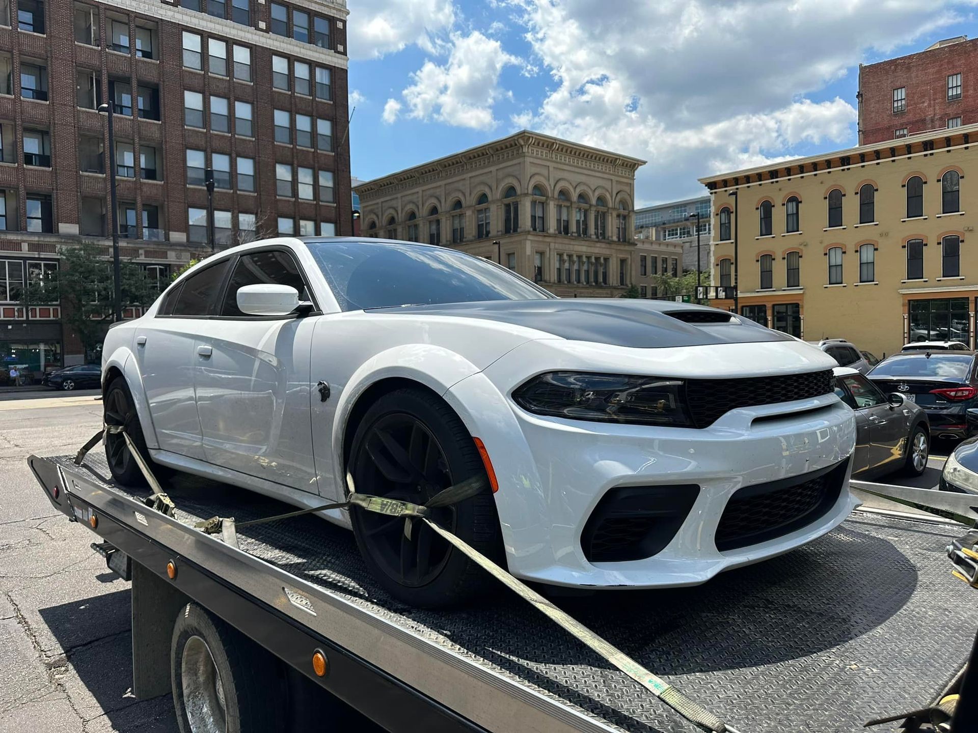 White Dodge Charger on a tow truck, black hood, sunny cityscape in background.