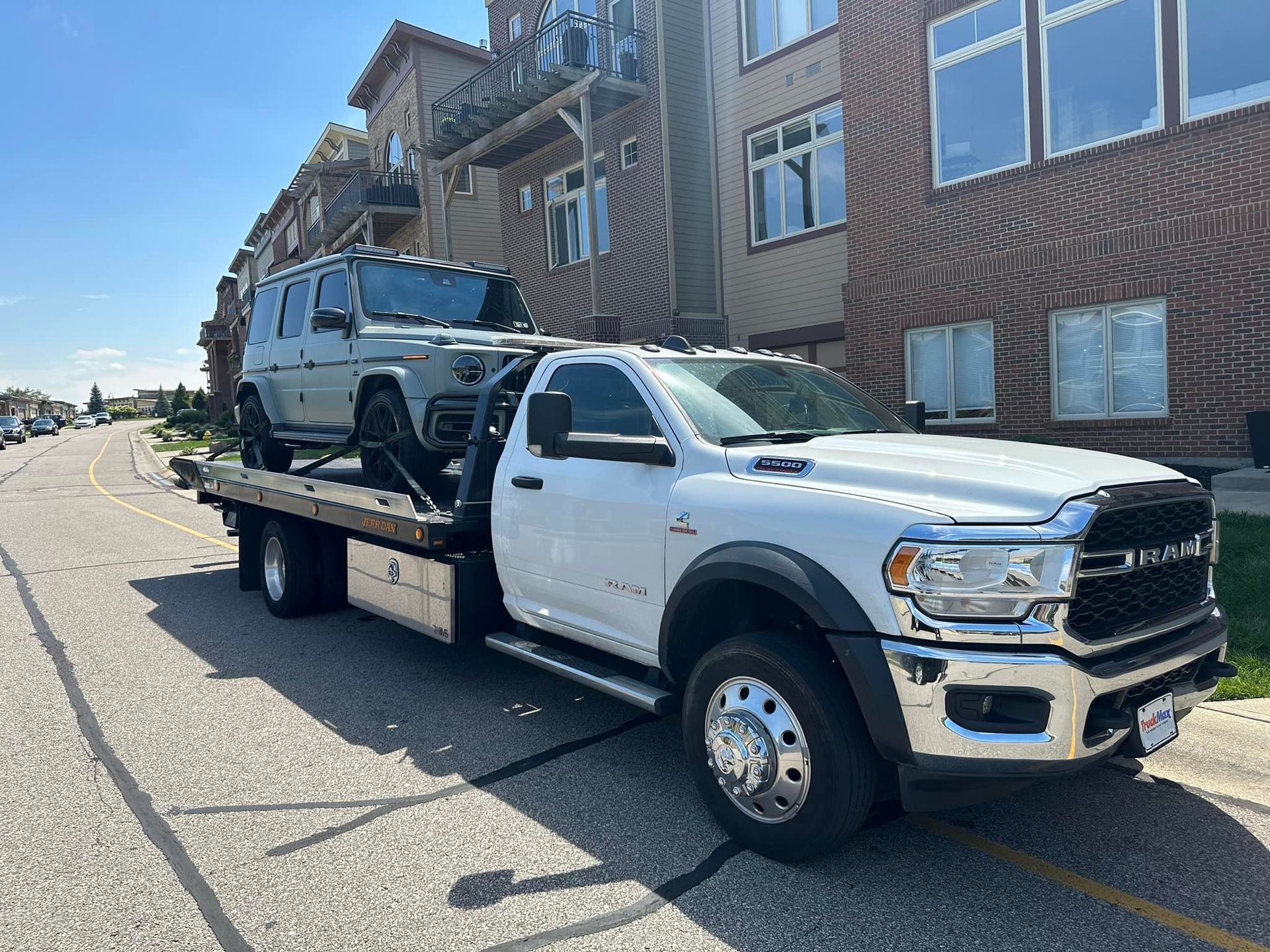 White tow truck carrying a gray Mercedes-Benz SUV on a city street in front of a brick building.