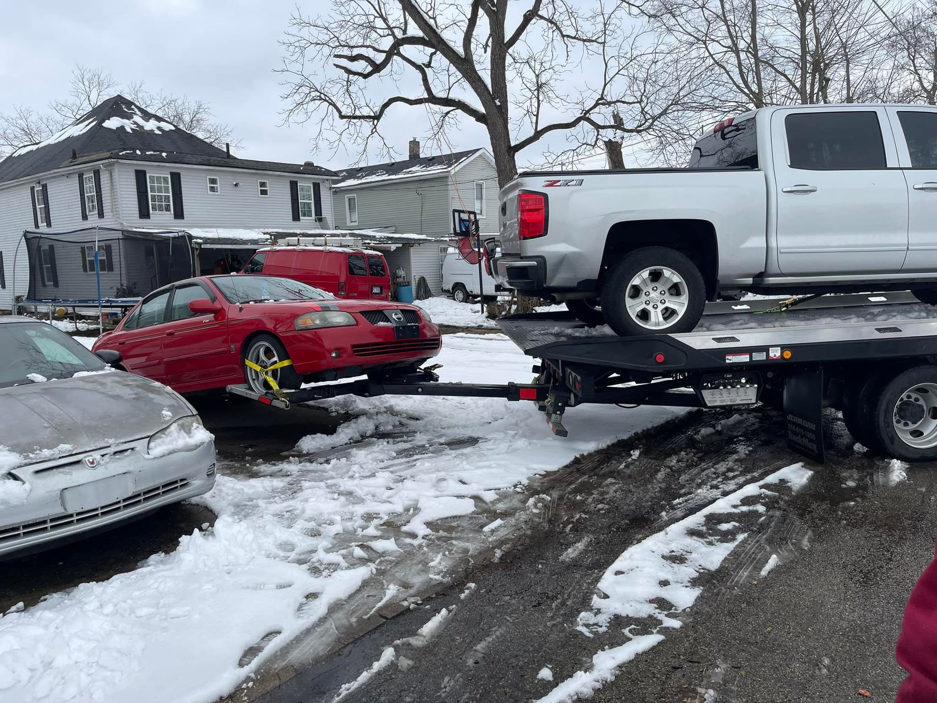 A red car on a tow truck being hauled by a silver pickup truck on a snow-covered street.