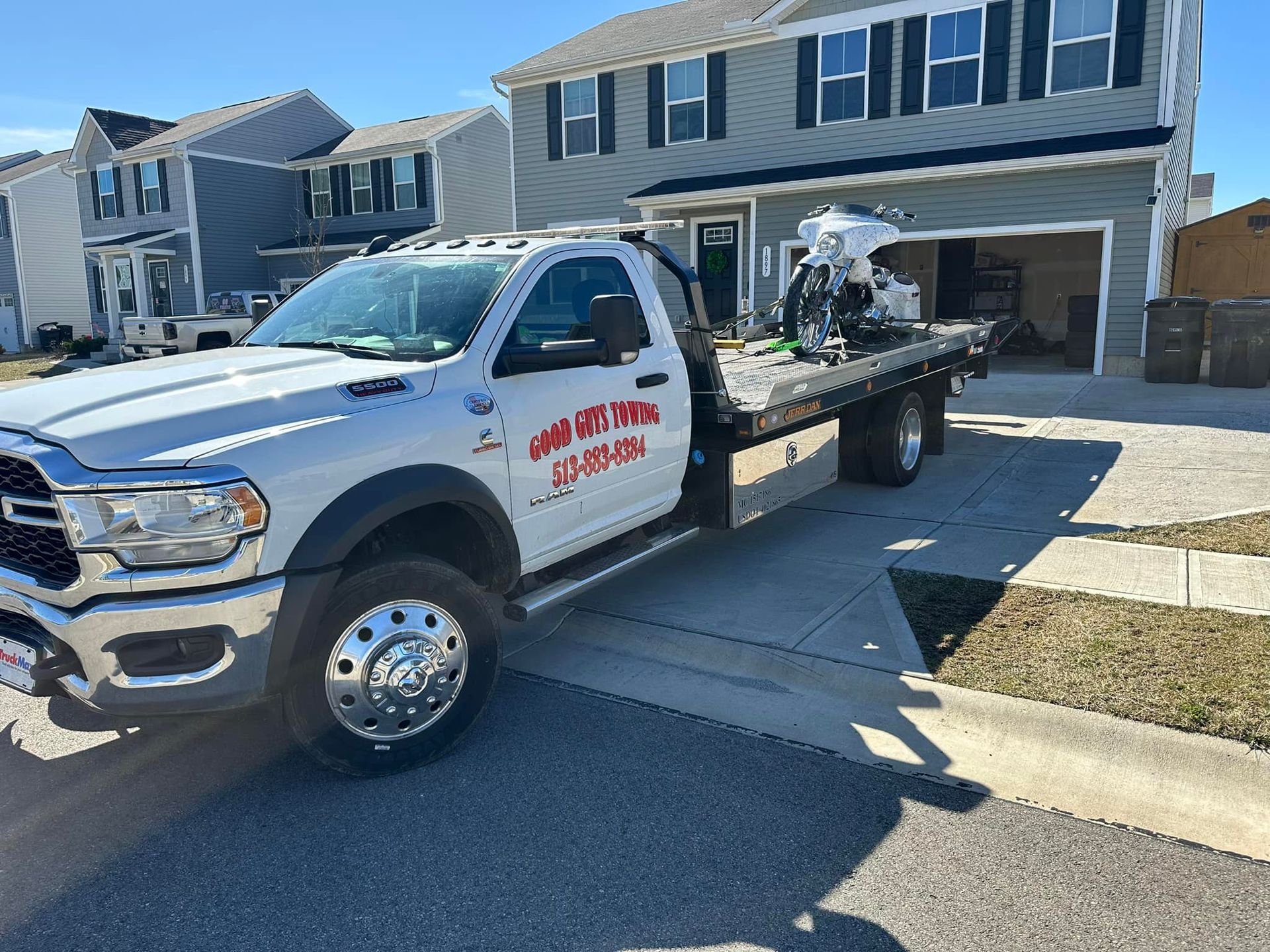 White tow truck parked in front of a house, carrying a motorcycle on its flatbed.