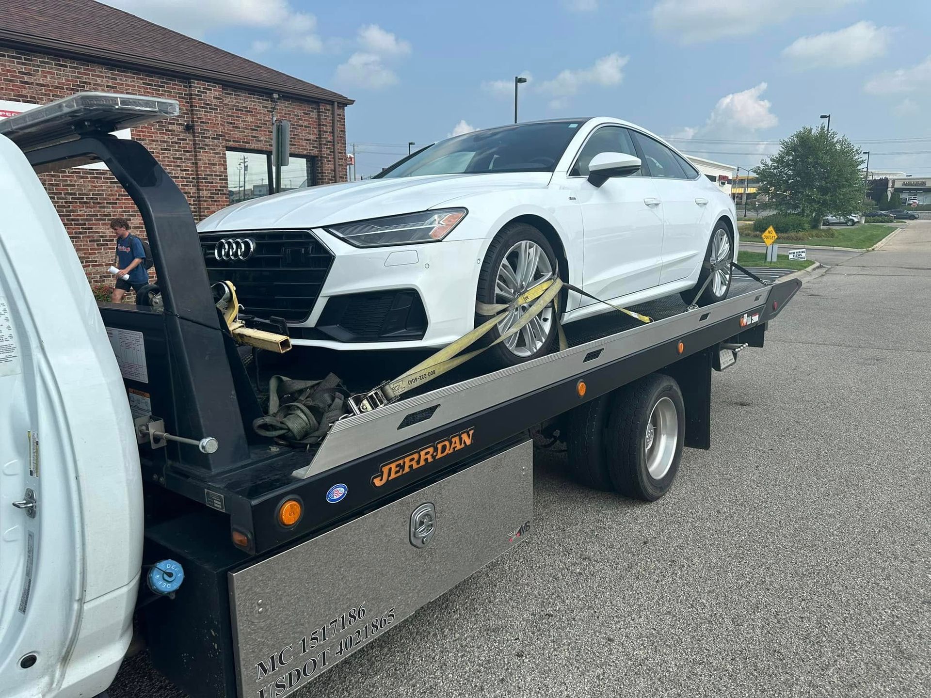 White Audi sedan being towed on a flatbed tow truck, outdoors near a brick building.