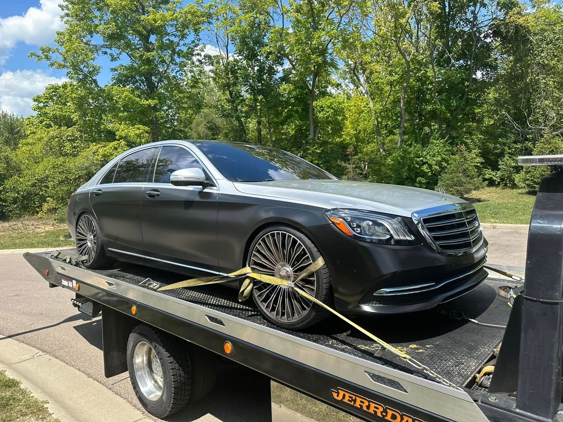 Silver and black Mercedes-Benz sedan on a tow truck, secured with straps, outdoors with trees in the background.