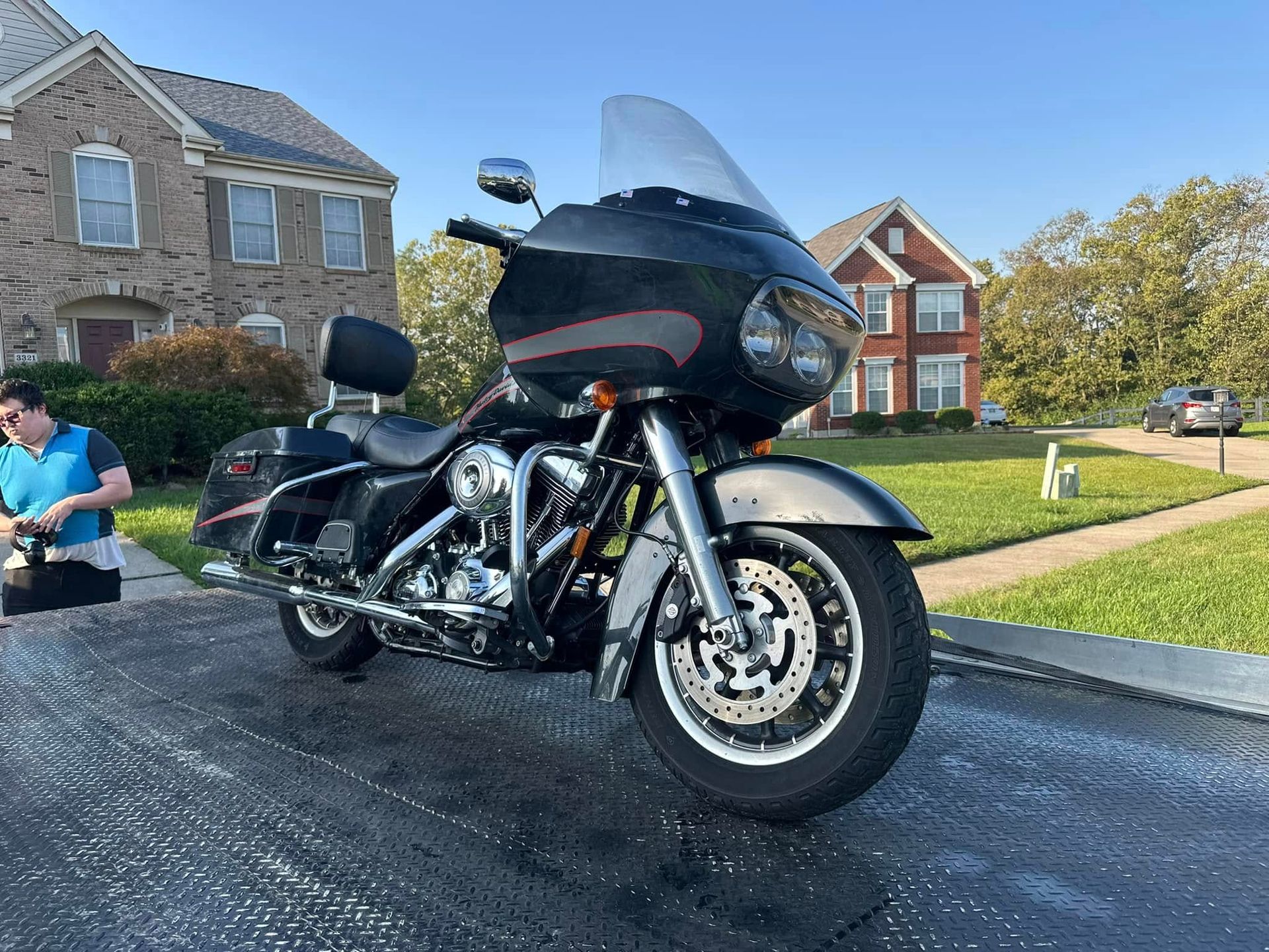 Black Harley Davidson motorcycle parked on a driveway with a person nearby. Houses and a blue sky are in the background.