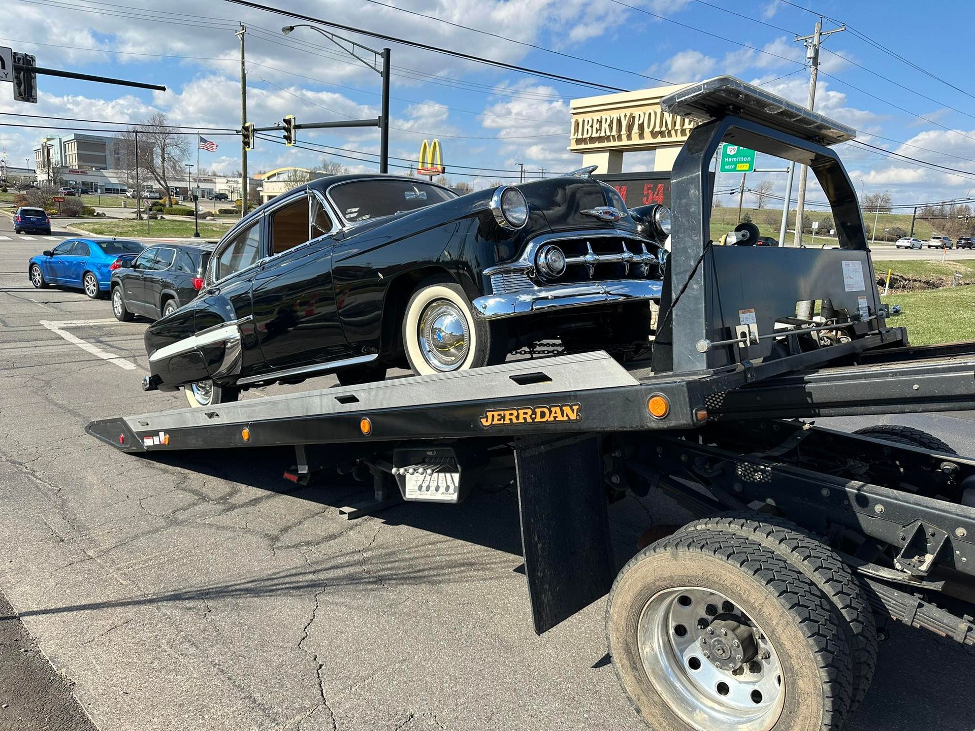 Black classic car on a tow truck at an intersection, damaged front, white tires, sunny day.