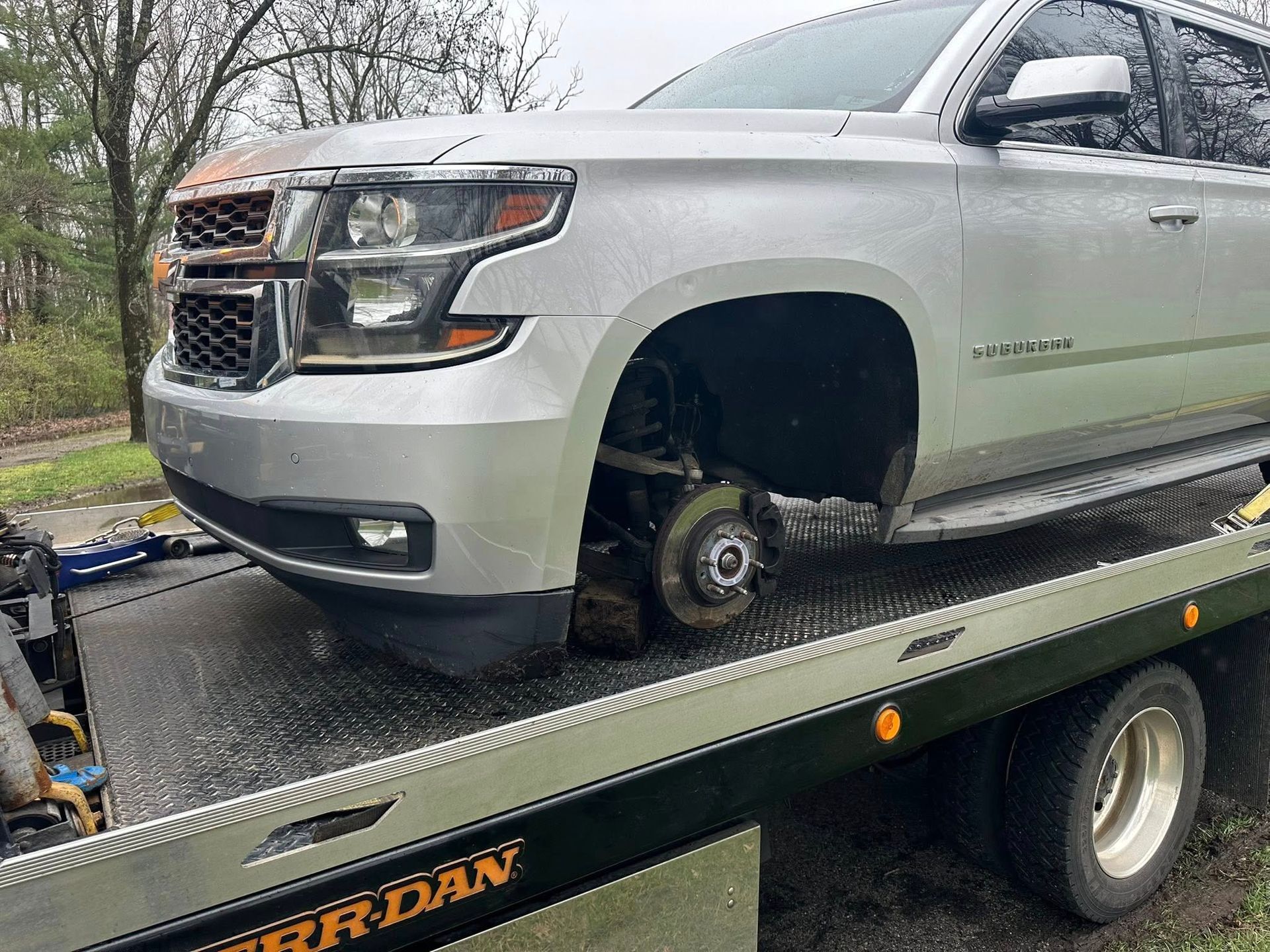 Silver SUV on a tow truck, front tire missing. Gray cloudy day.