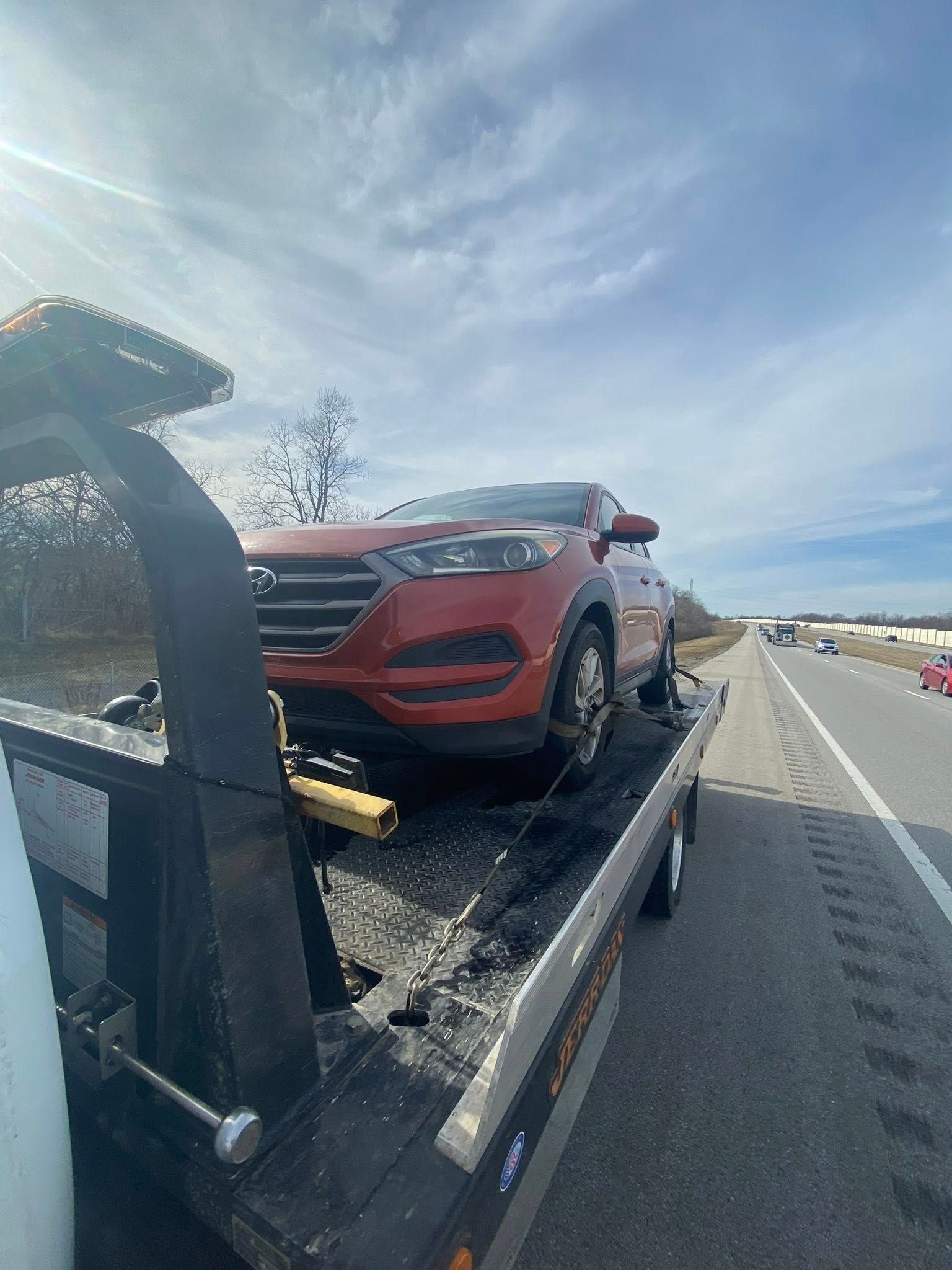 Red car being towed on a flatbed tow truck on a highway under a cloudy sky.
