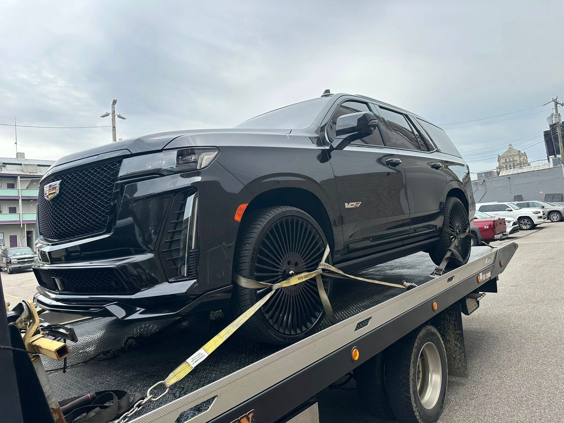 Black Cadillac Escalade SUV on a tow truck, secured with straps, parked outdoors.