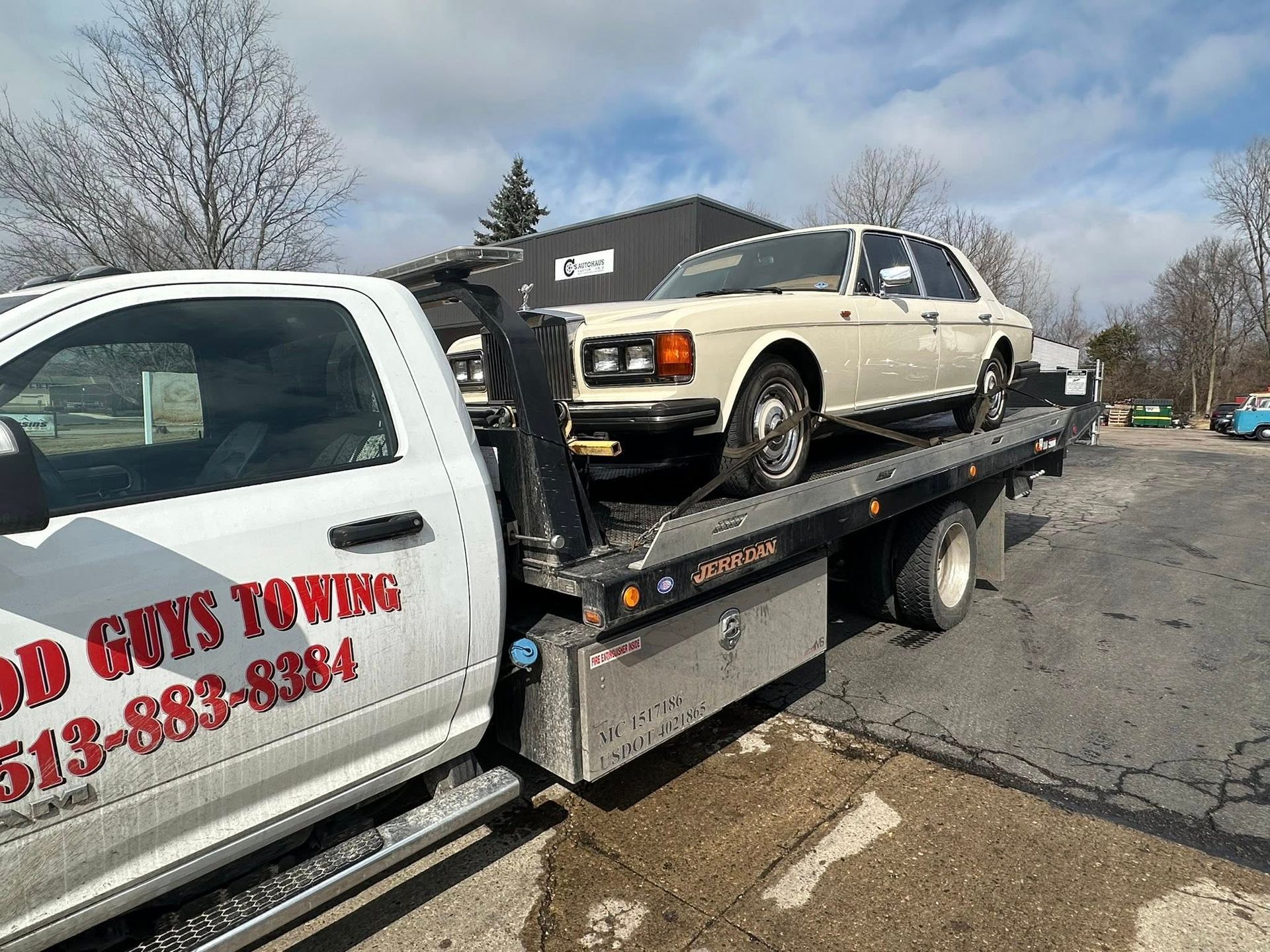 Tow truck with a cream-colored vintage car on its flatbed; Good Guys Towing logo on truck. Outdoors.