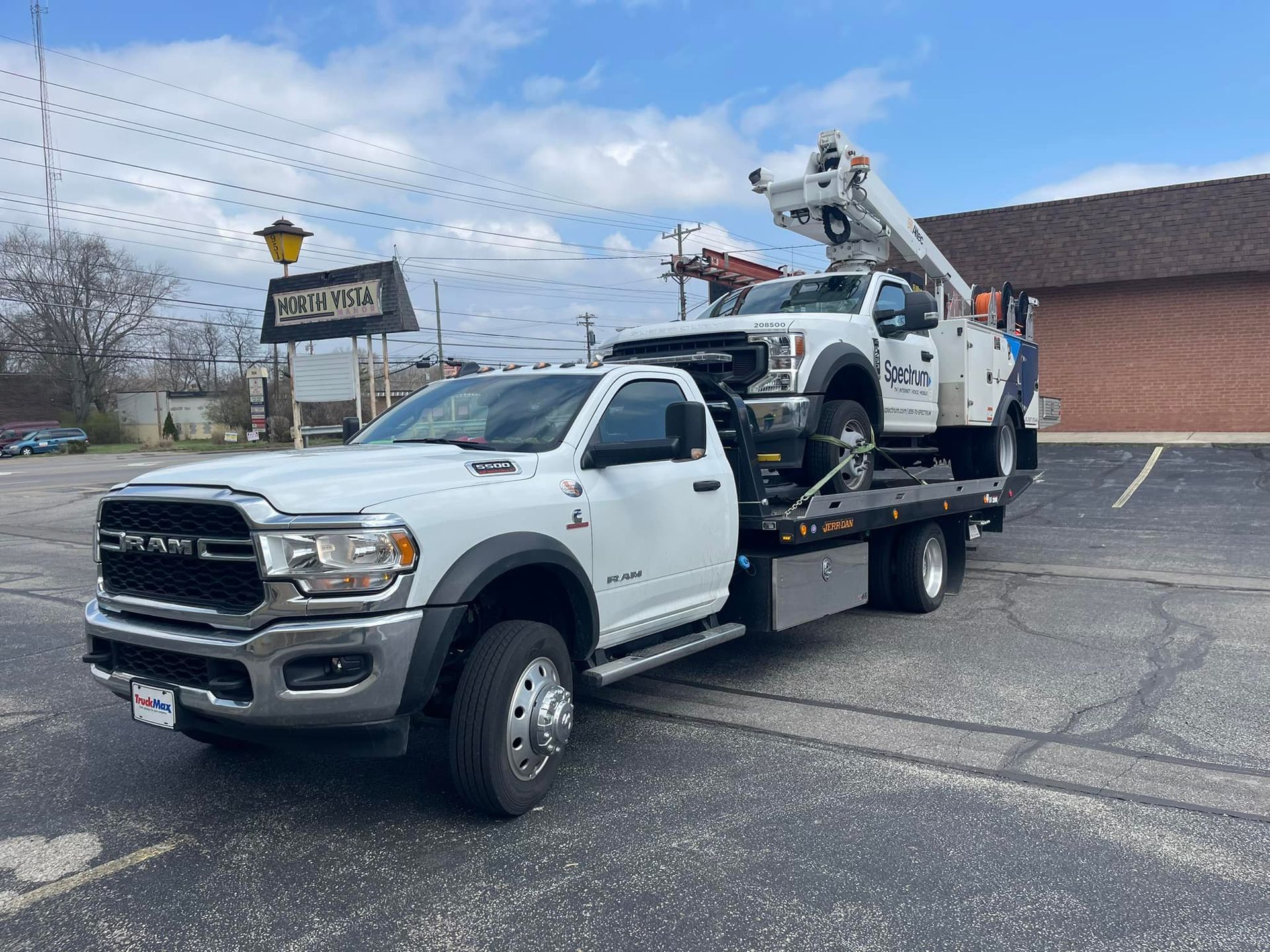 White tow truck carrying a white utility truck with raised bucket, parked in a lot.