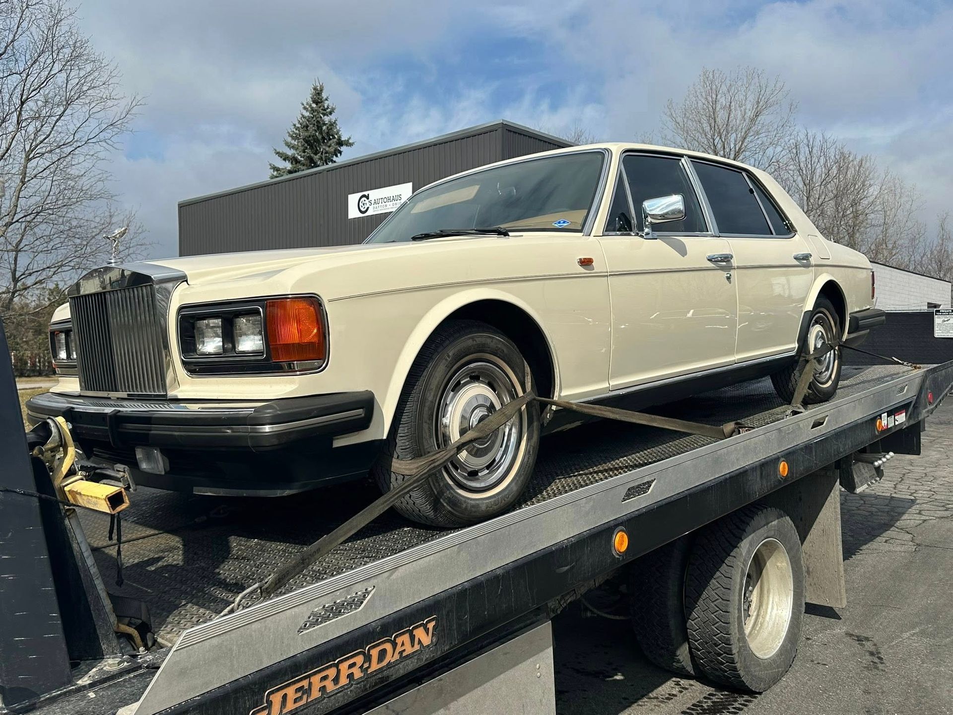 Cream-colored Rolls-Royce sedan on a tow truck, secured with straps. It's parked outside a building on a sunny day.