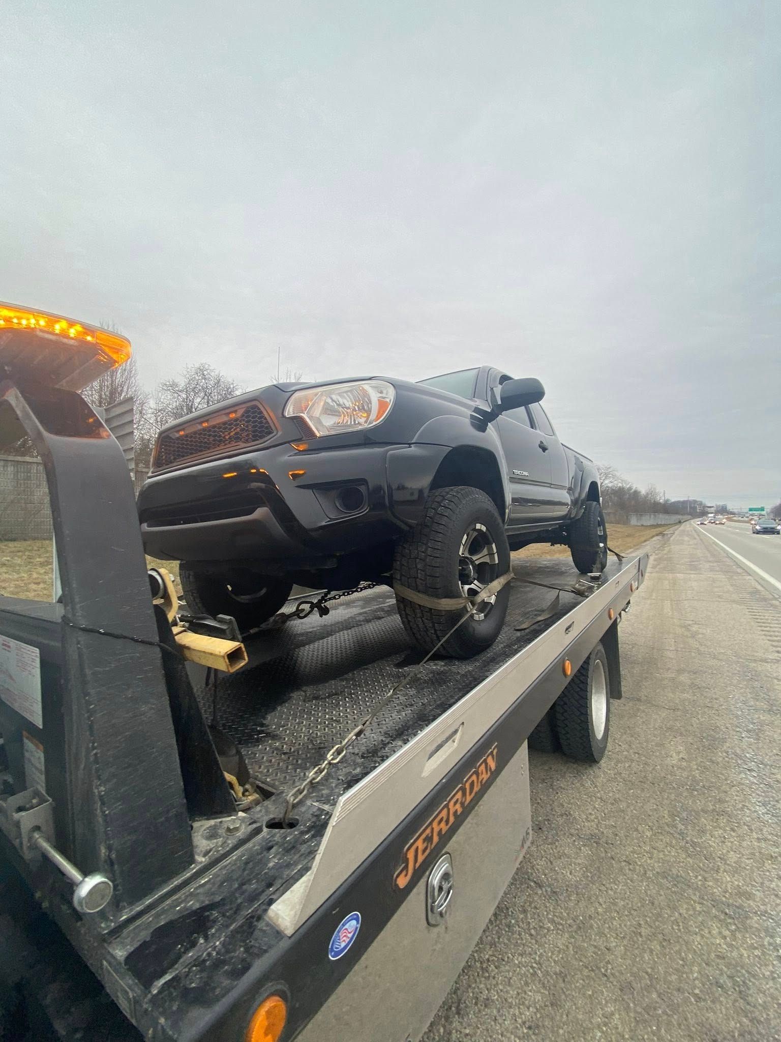 Black truck on a tow truck on the side of a road under a cloudy sky.