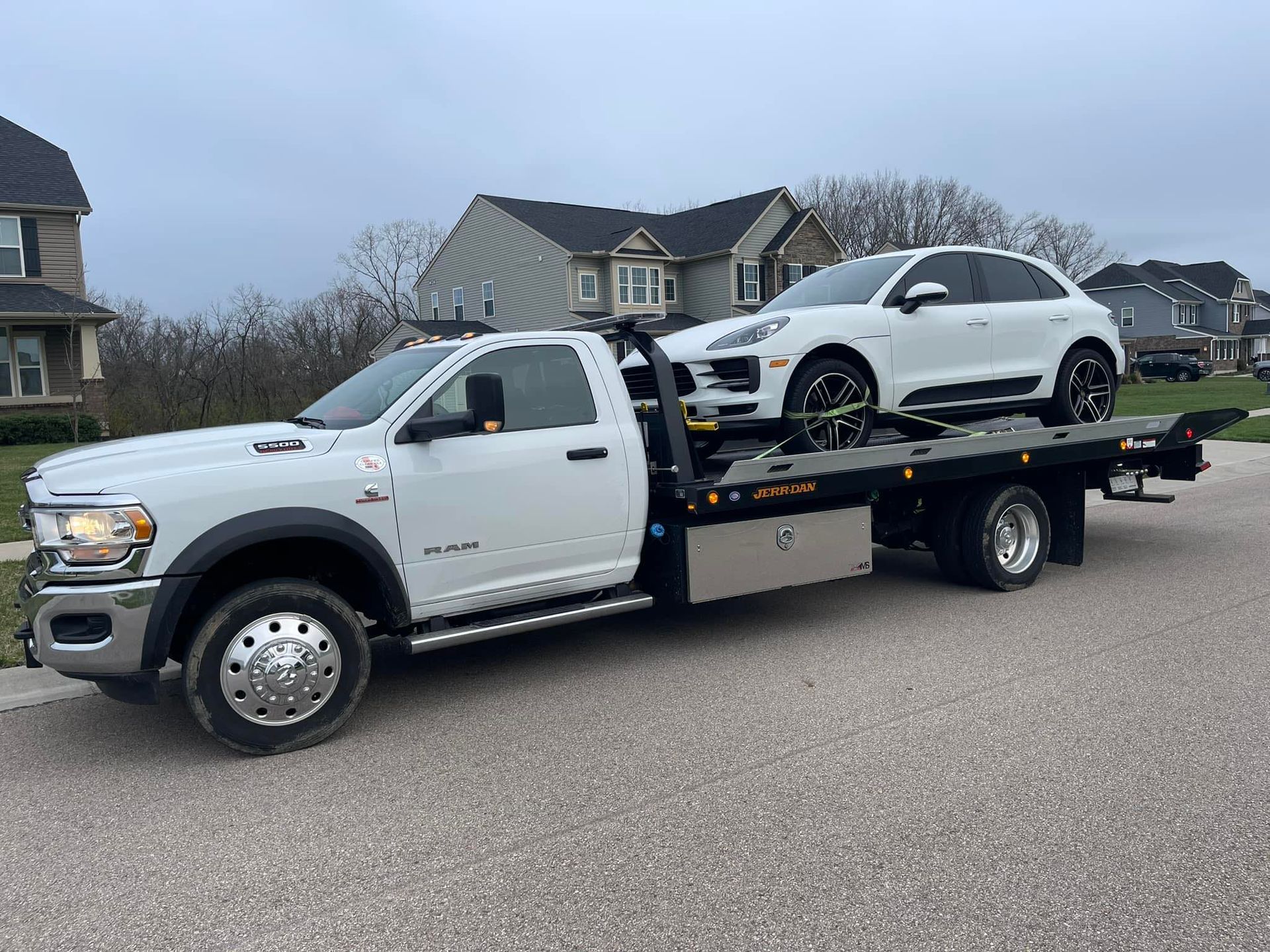 White tow truck carrying a white Porsche SUV on a residential street. Cloudy sky.