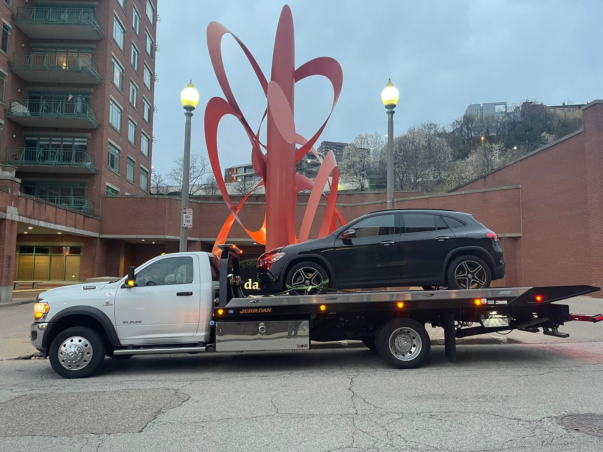 A black SUV on a flatbed tow truck in front of a red sculpture and brick building.