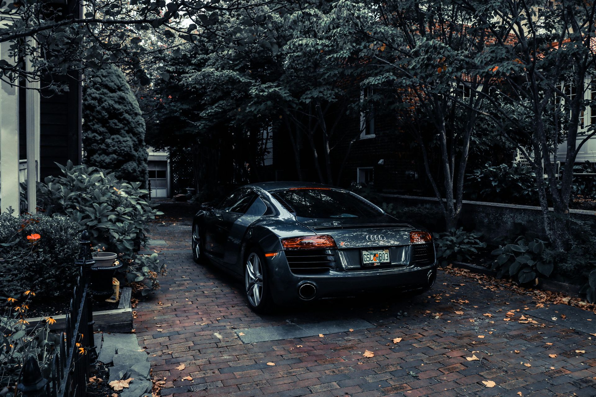 Dark grey Audi R8 parked on a brick driveway lined with bushes and trees.
