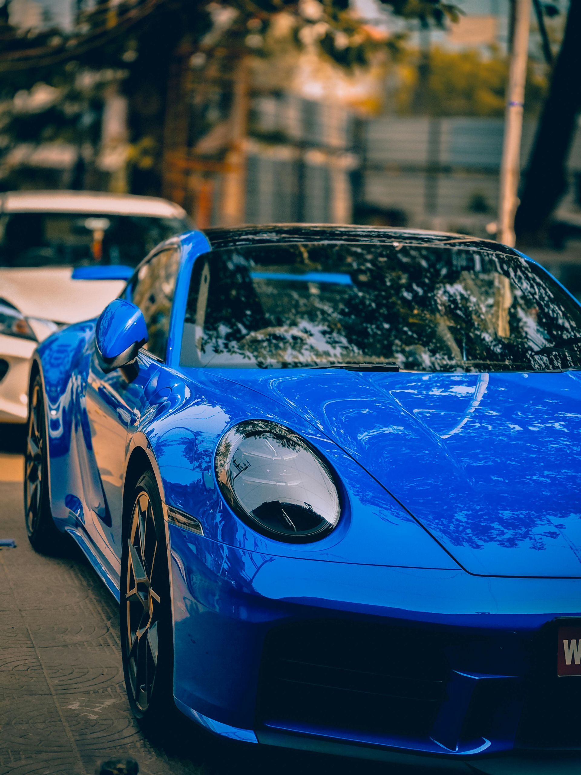 Blue sports car parked on a street, with a white car partially visible in the background.