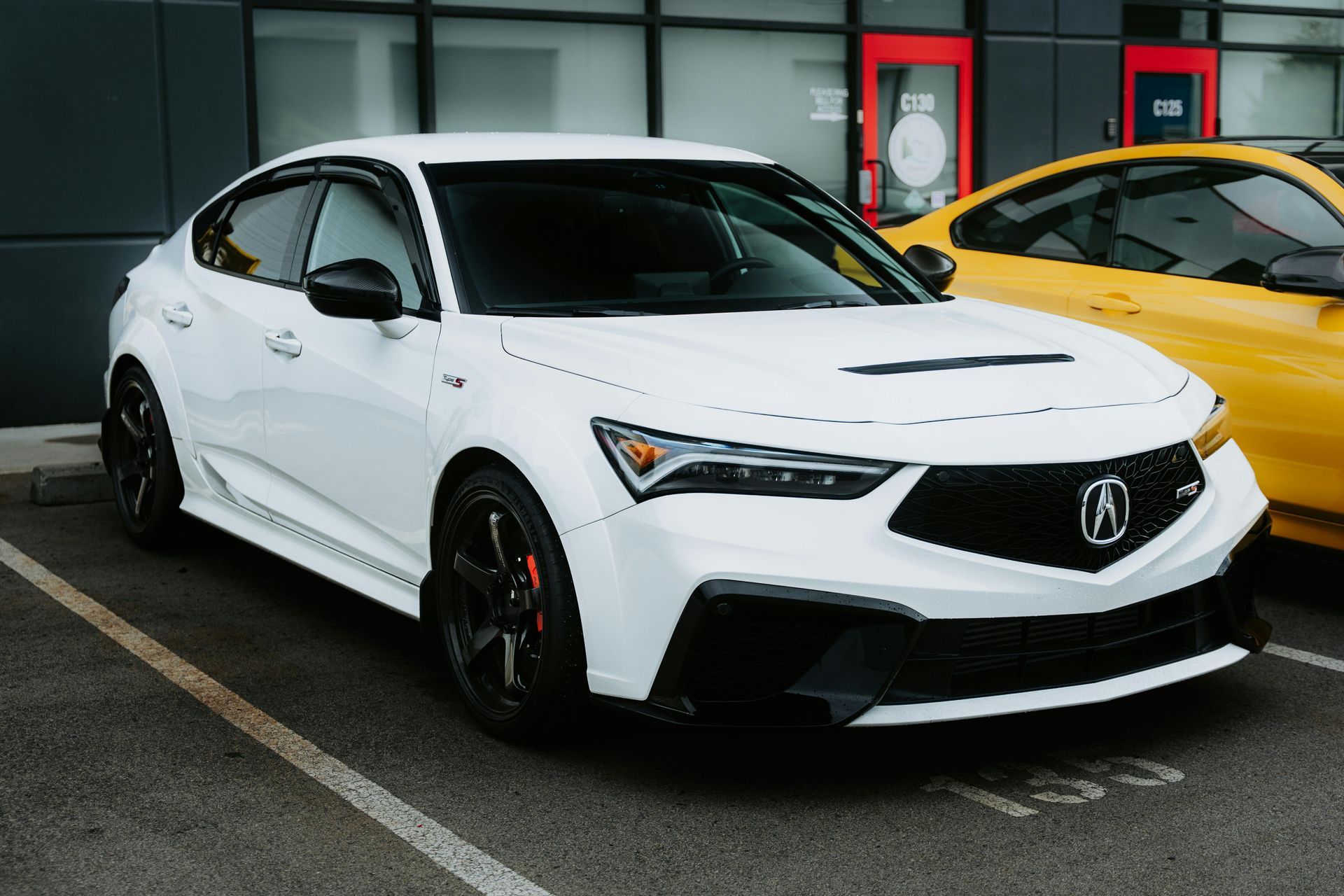 White Acura Integra parked in front of a building, black accents, red brake calipers, next to a yellow car.