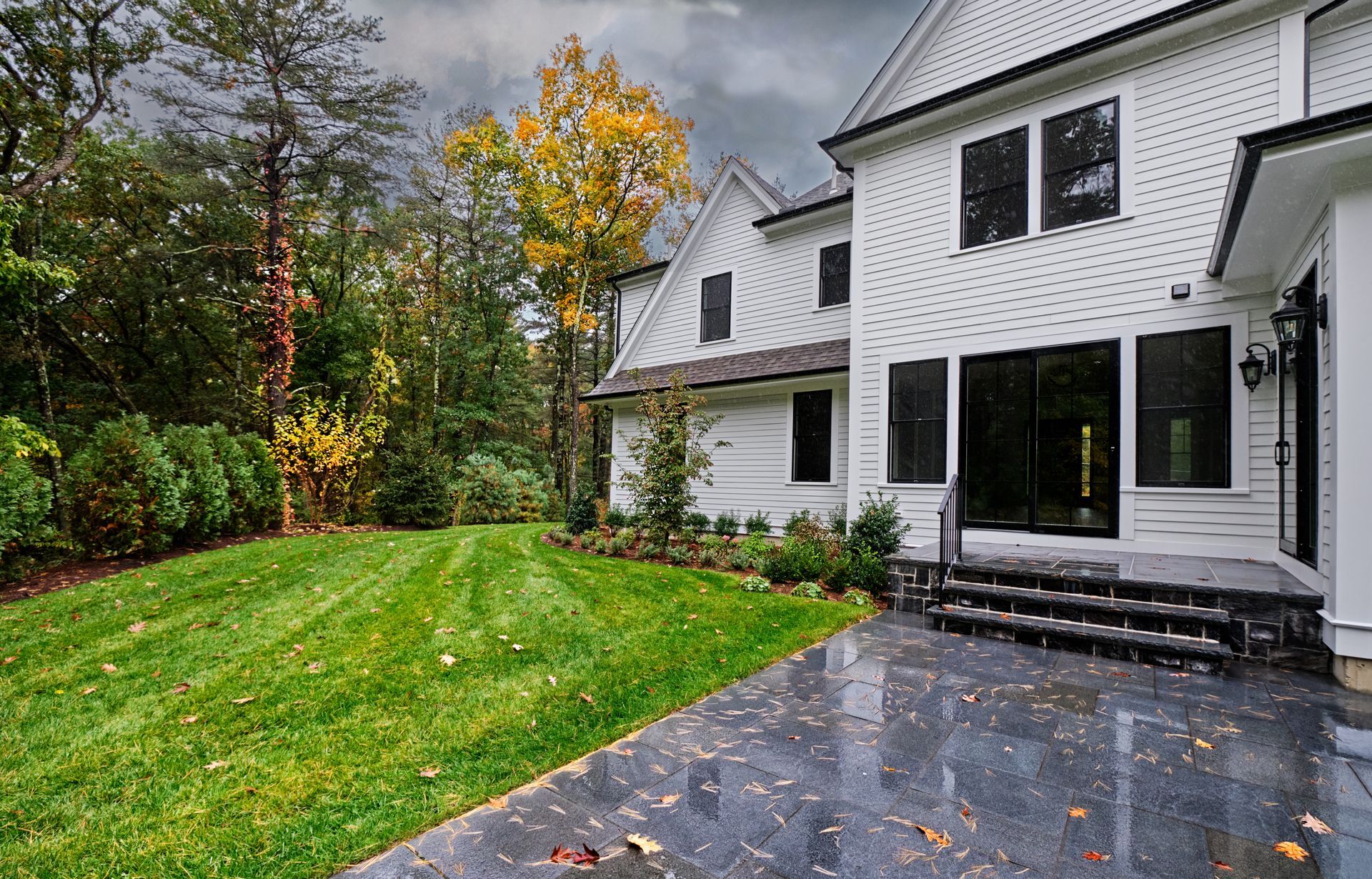 Maison blanche aux fenêtres sombres, terrasse grise et pelouse verte devant. On aperçoit des feuilles d'automne et des arbres.