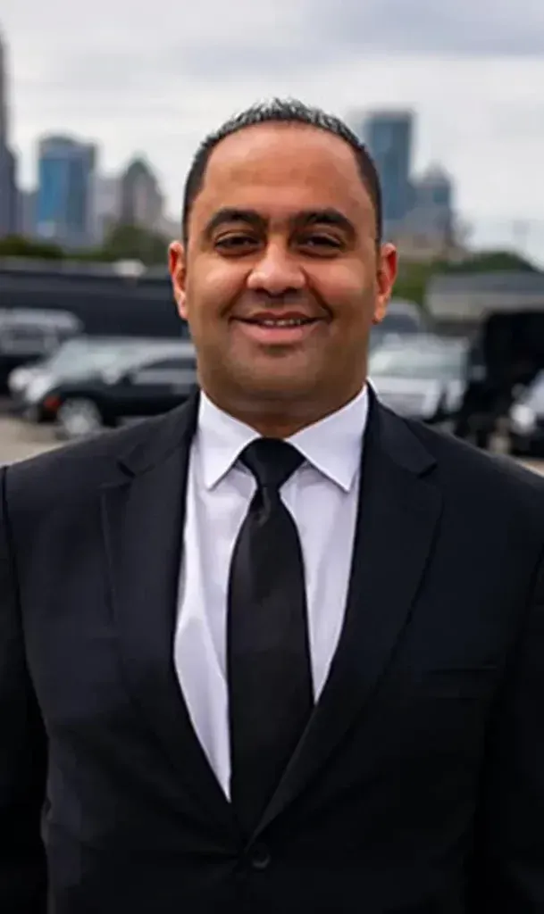 Man in black suit and tie smiles, with a city skyline and cars blurred in the background.