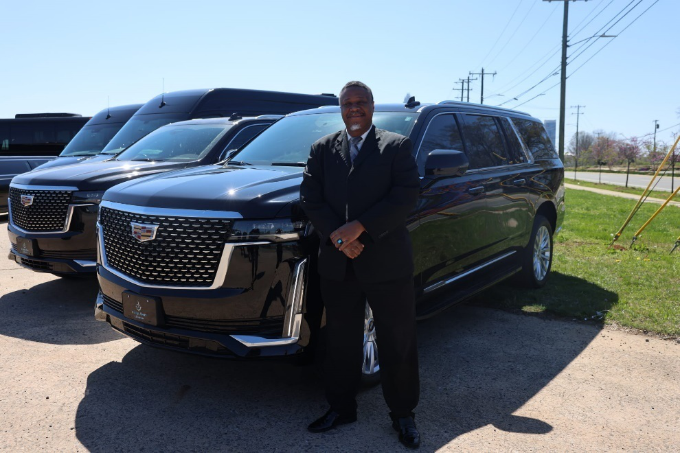 Man in black suit standing by black Cadillac SUVs on a sunny day.