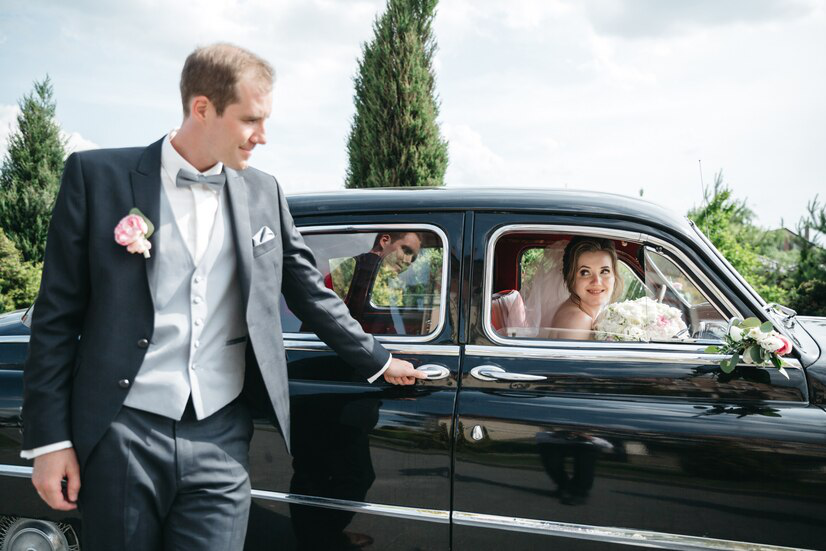 Groom opens car door for bride; they smile. Black vintage car, sunny day.