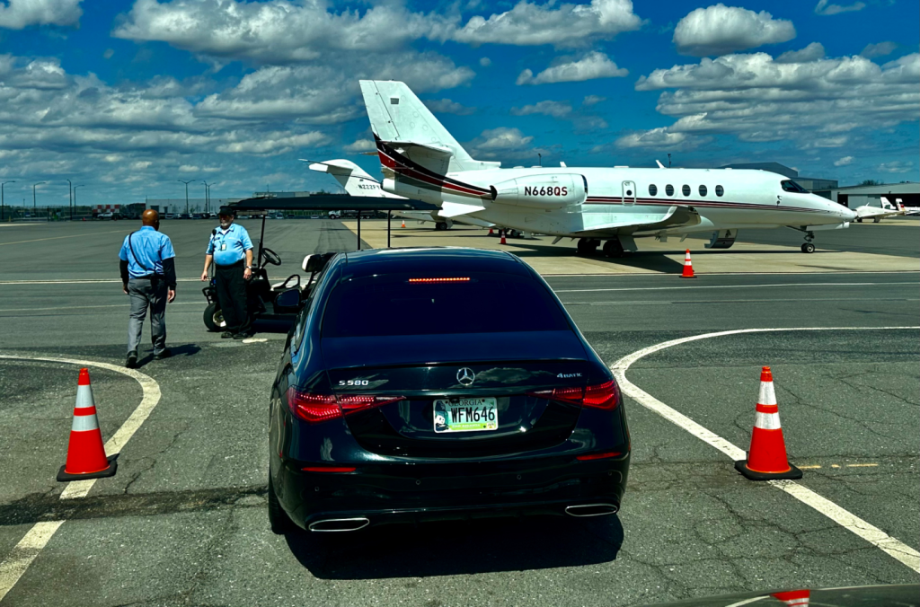 A black car on tarmac with two security officers nearby and a private jet in the background.